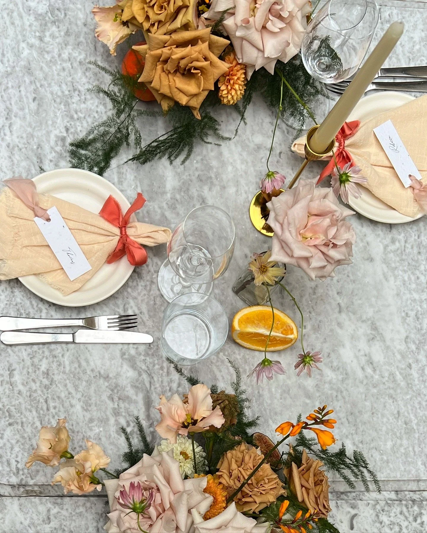 Overhead view of a table set for a meal with floral arrangements, two place settings with folded napkins and ribbons, glasses, cutlery, a sliced orange, and a gold candle holder with a candle.