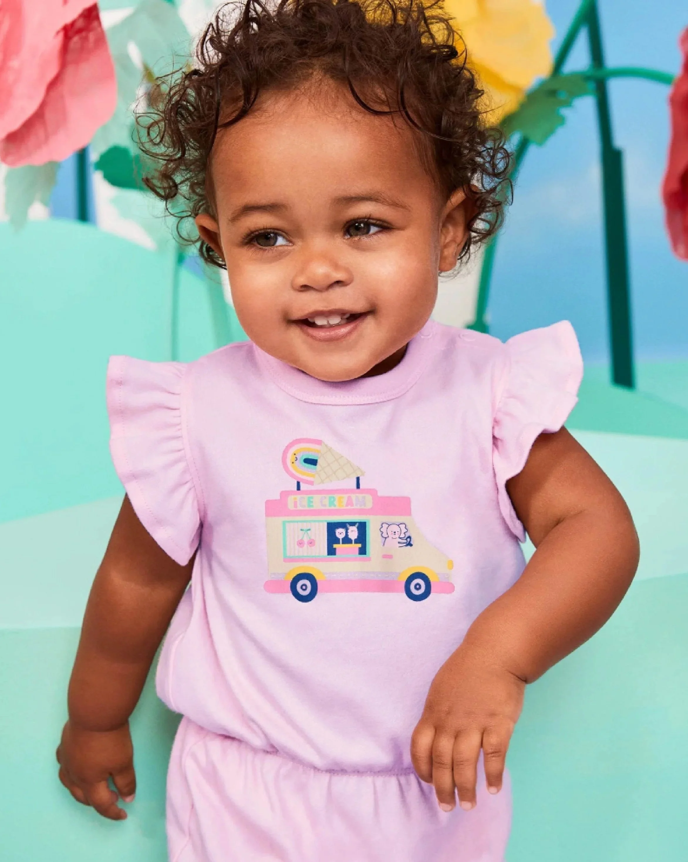 Close-up of a young girl with dark curly hair, wearing a pink T-shirt with a colorful ice cream truck illustration. She is smiling and looking to the side, with a playful expression. Background appears to be a pastel-colored setting.