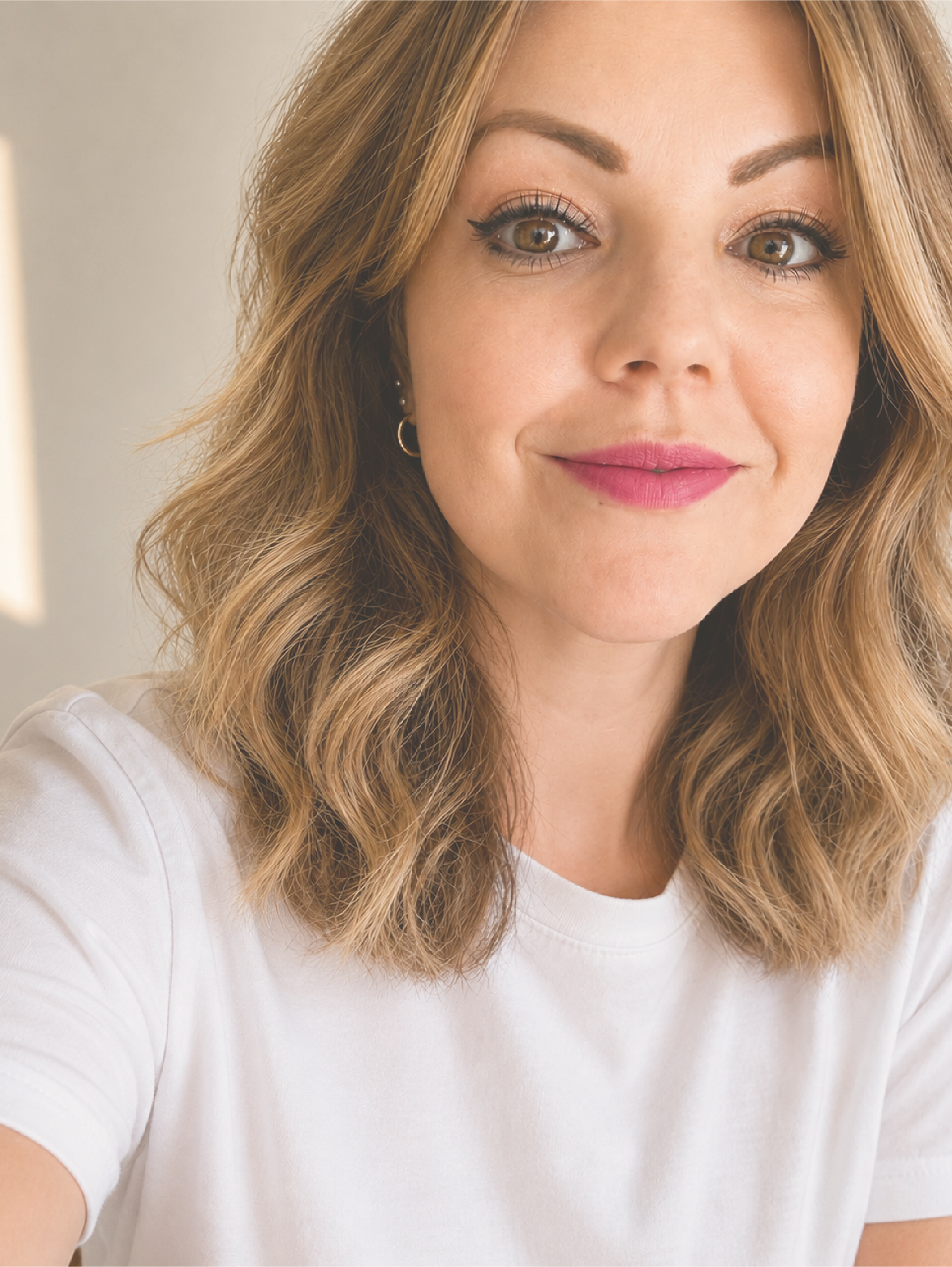 Close-up of a woman with wavy, light brown hair, wearing makeup with eyeliner and lipstick, and a white t-shirt.
