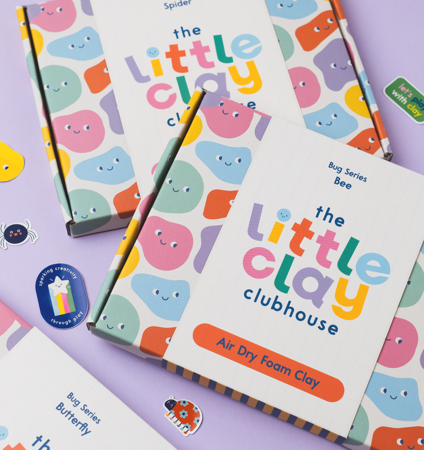 Colorful themed children's educational clay modeling kit titled "The Little Clay Clubhouse" with packaging featuring smiling, pastel-colored cloud characters, along with stickers of bugs and a rainbow star.