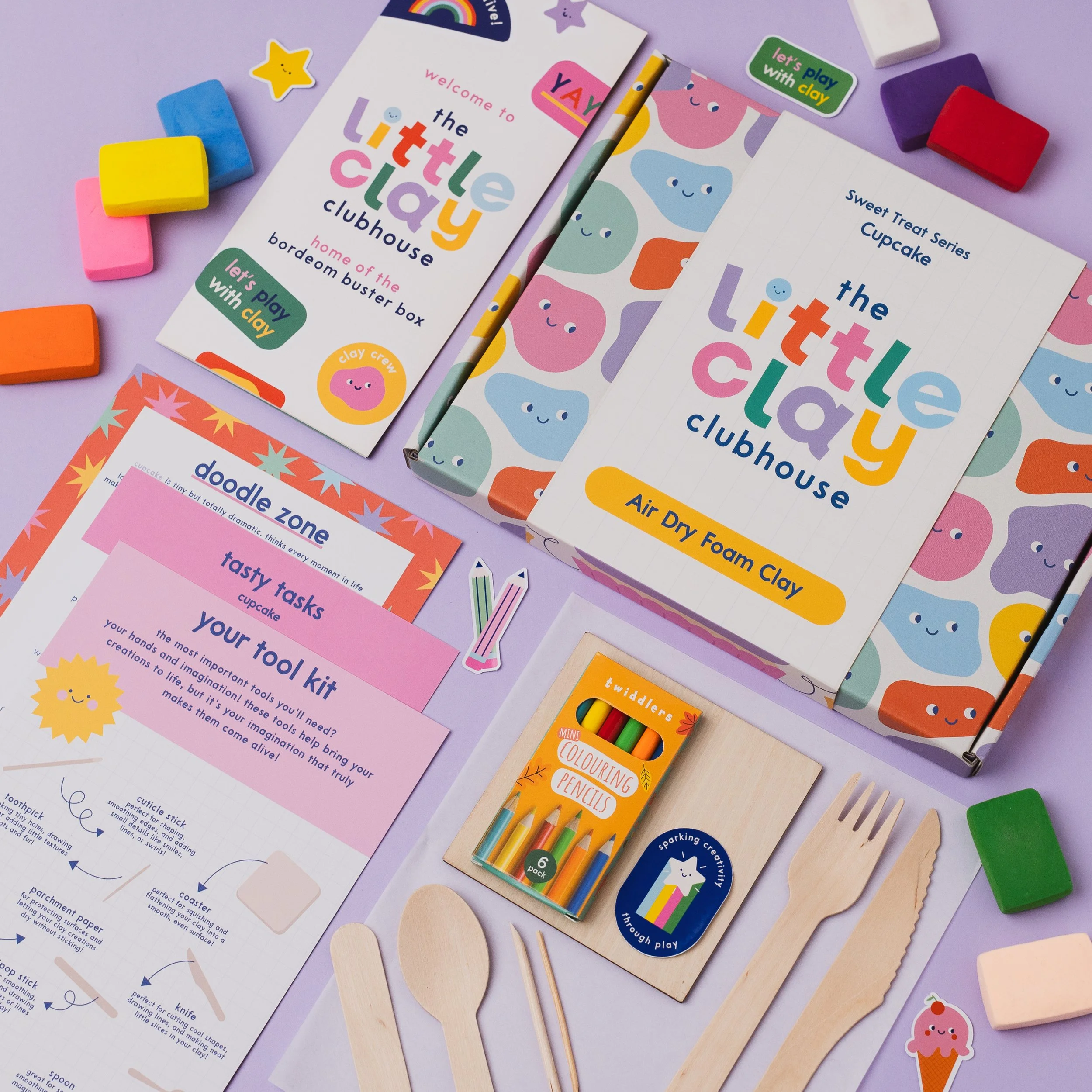 A colorful assortment of children's arts and crafts supplies on a light purple background, including wooden spoons, blocks, paper, stickers, and a magazine with playful graphics and text related to clay modeling activities.