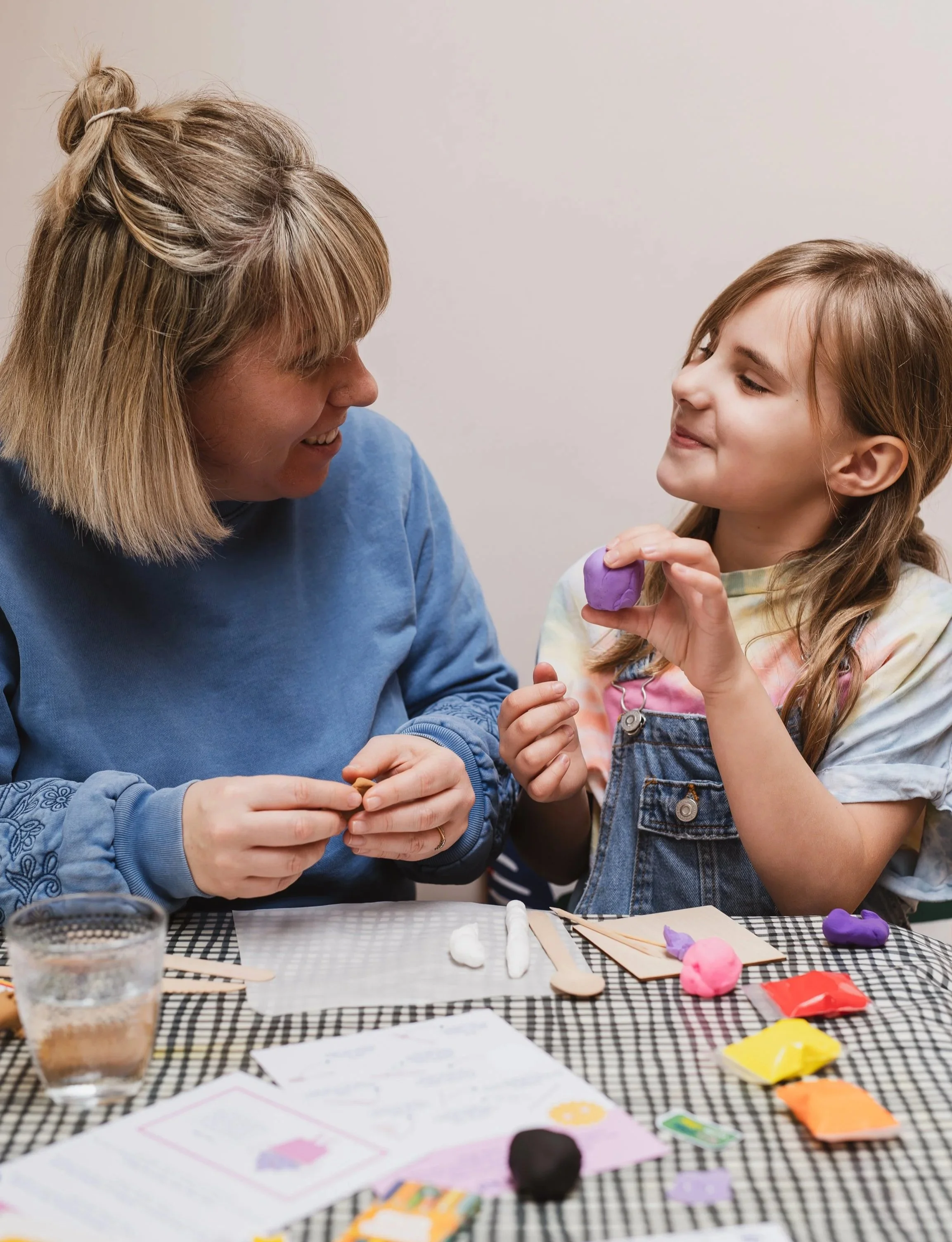 A woman and a girl sitting at a table, engaging in a crafting activity with colorful modeling clay and paper, with a glass of water and craft supplies in the foreground.