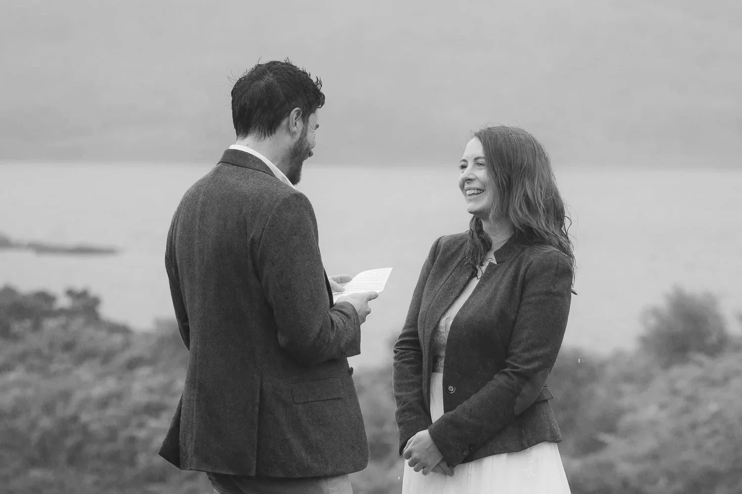 Black and white photo of groom reading vows, smiling as raindrops fall