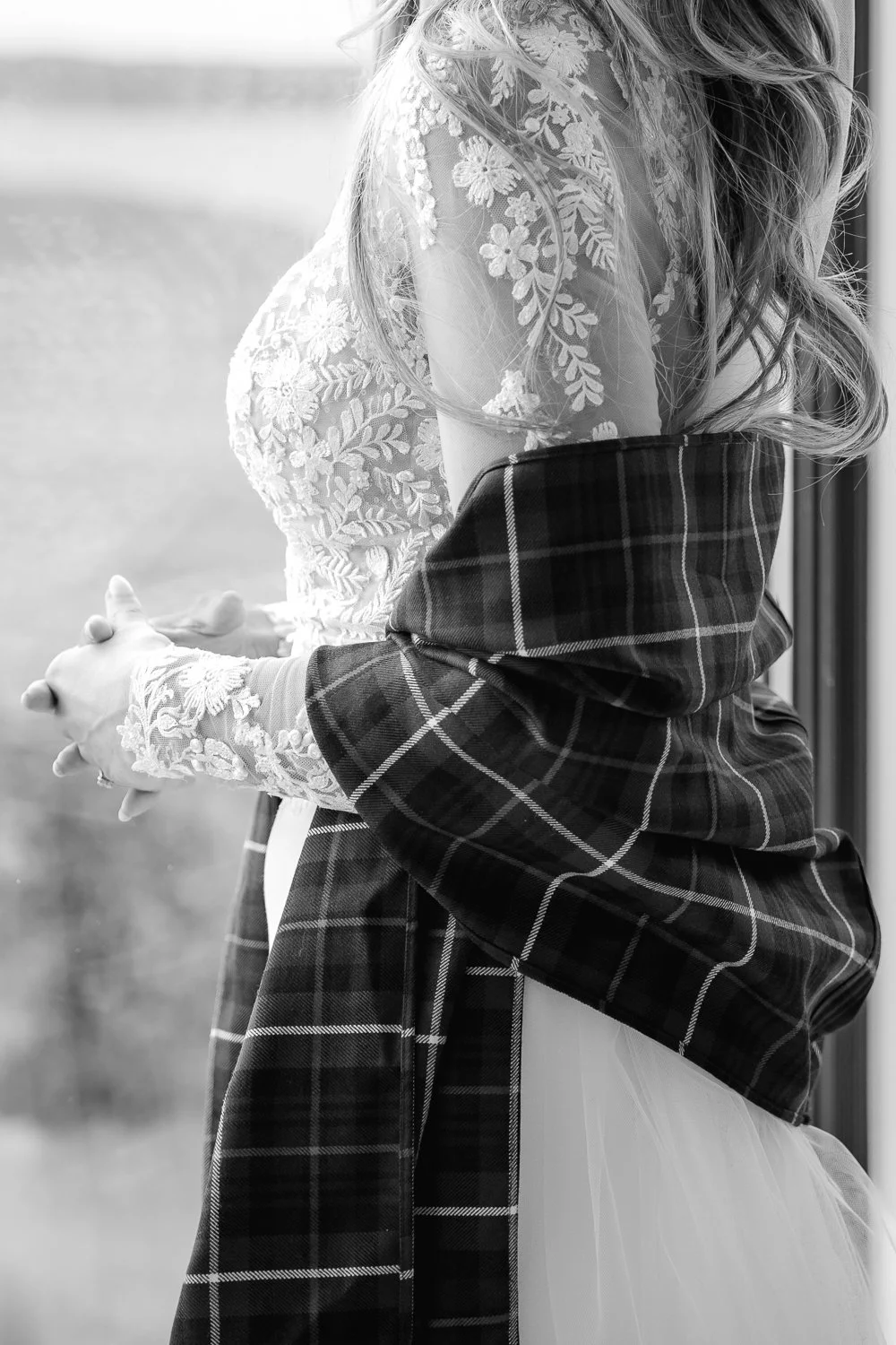 Black and white close-up of a bride in a lace wedding dress, draped in a traditional Scottish tartan shawl, standing by a window.