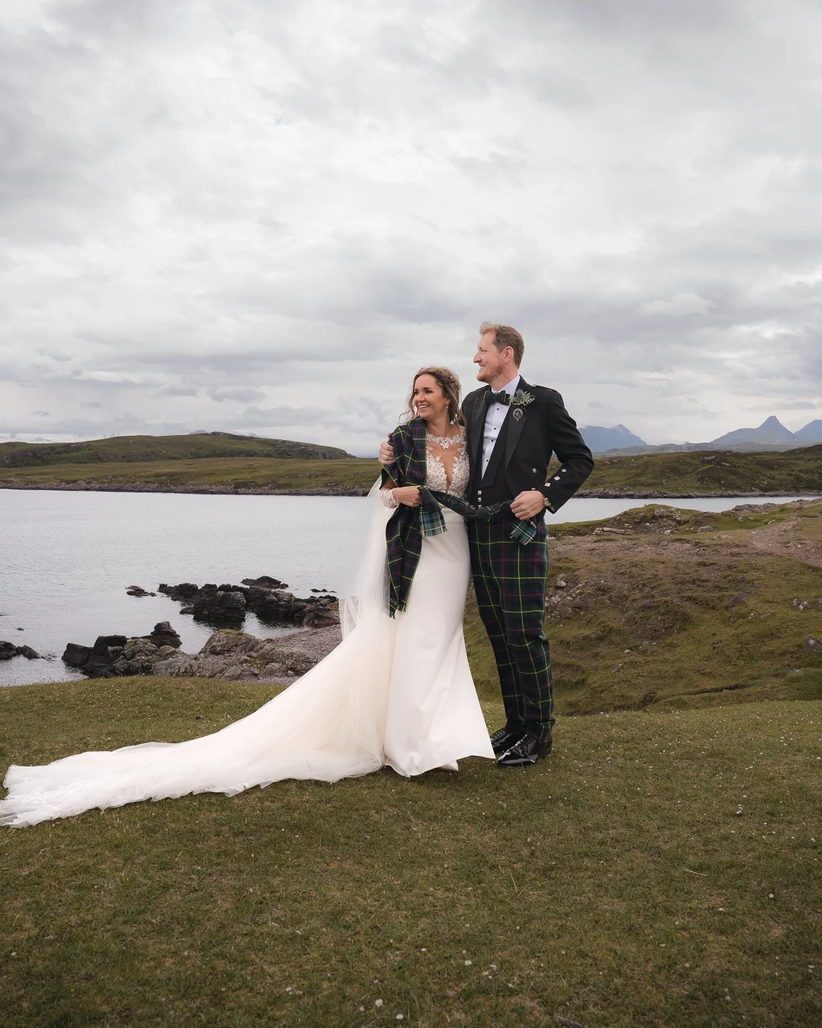 Bride and groom in traditional Scottish wedding attire embracing by a loch in the Highlands, with dramatic mountains and moody skies in the background.