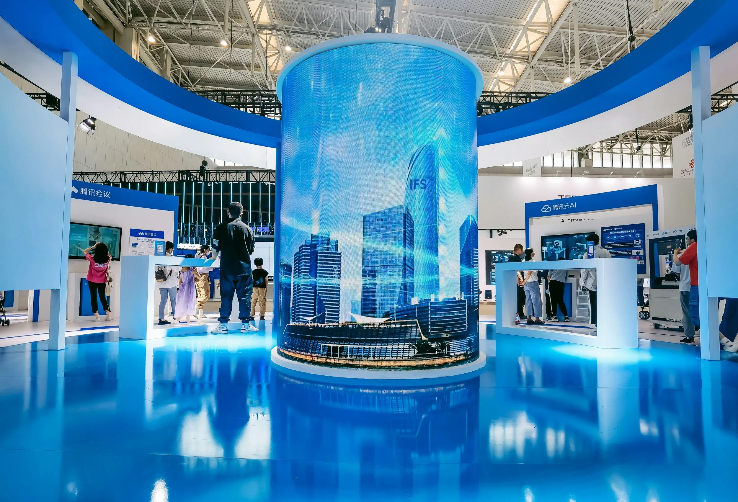 A high-tech exhibition hall with a large cylindrical display showing a city skyline. People are exploring various electronic displays and kiosks around. The ceiling has a geometric design with large overhead lights