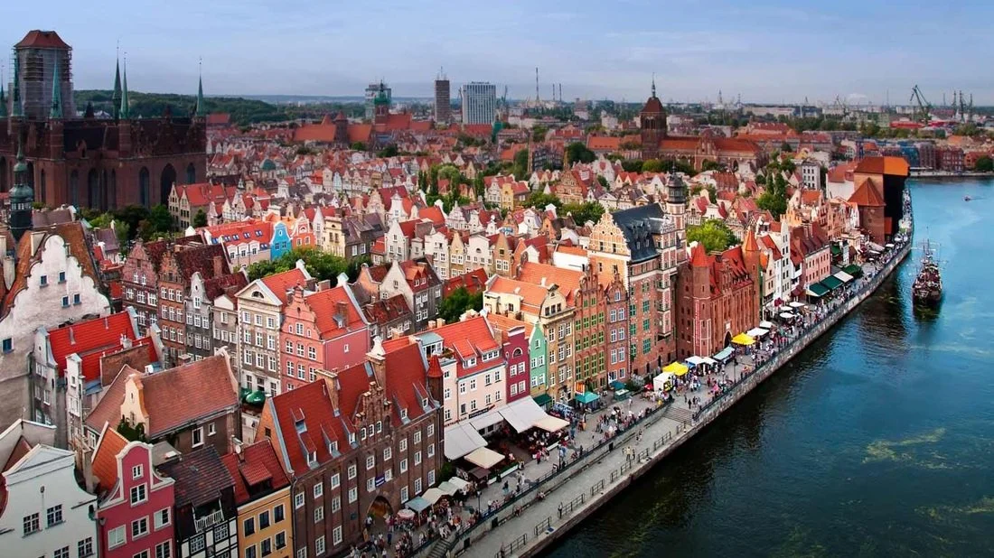 Aerial view of colorful waterfront city with boats, crowds, and a historic church in the background.
