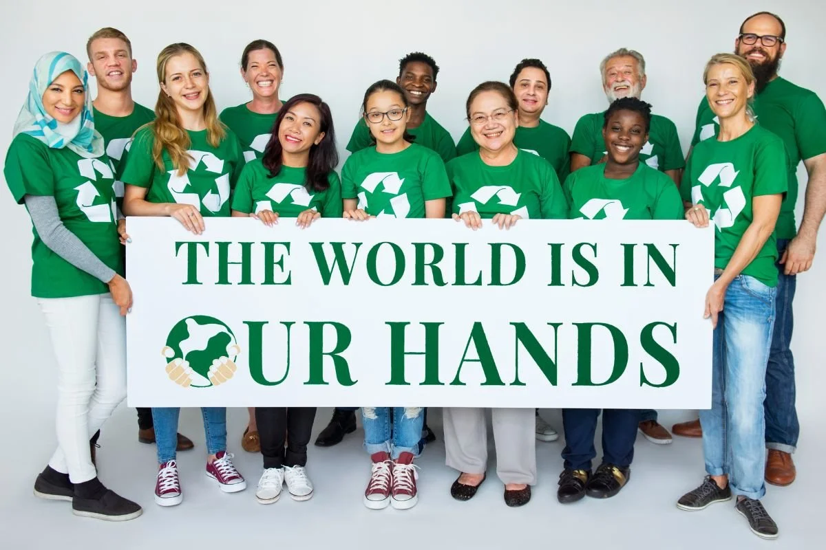 A diverse group of people wearing green shirts with recycling symbols hold a large sign that reads, THE WORLD IS IN OUR HANDS. They are smiling, standing together on a plain background.