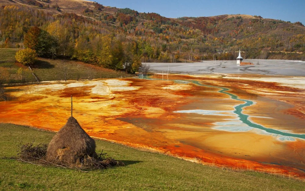A vibrant scene with a haystack in a grassy field, and a colorful landscape of orange, yellow, and blue hues from pollution. In the background, a church steeple emerges from a partially flooded area amidst forested hills under a clear sky.