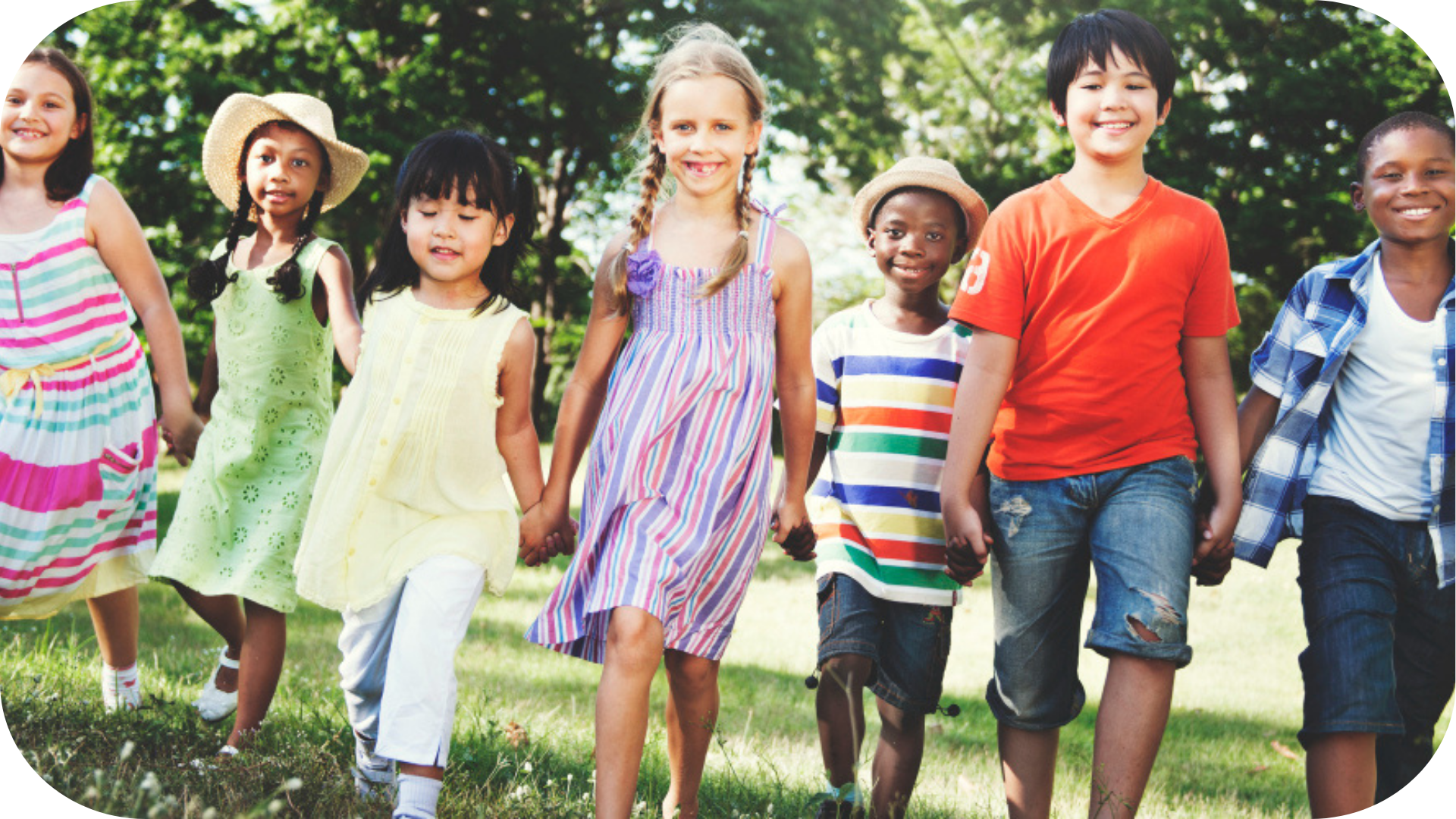 A diverse group of happy children holding hands and walking together outside on a sunny day. They are smiling, wearing colorful summer clothes, with trees and grass in the background.