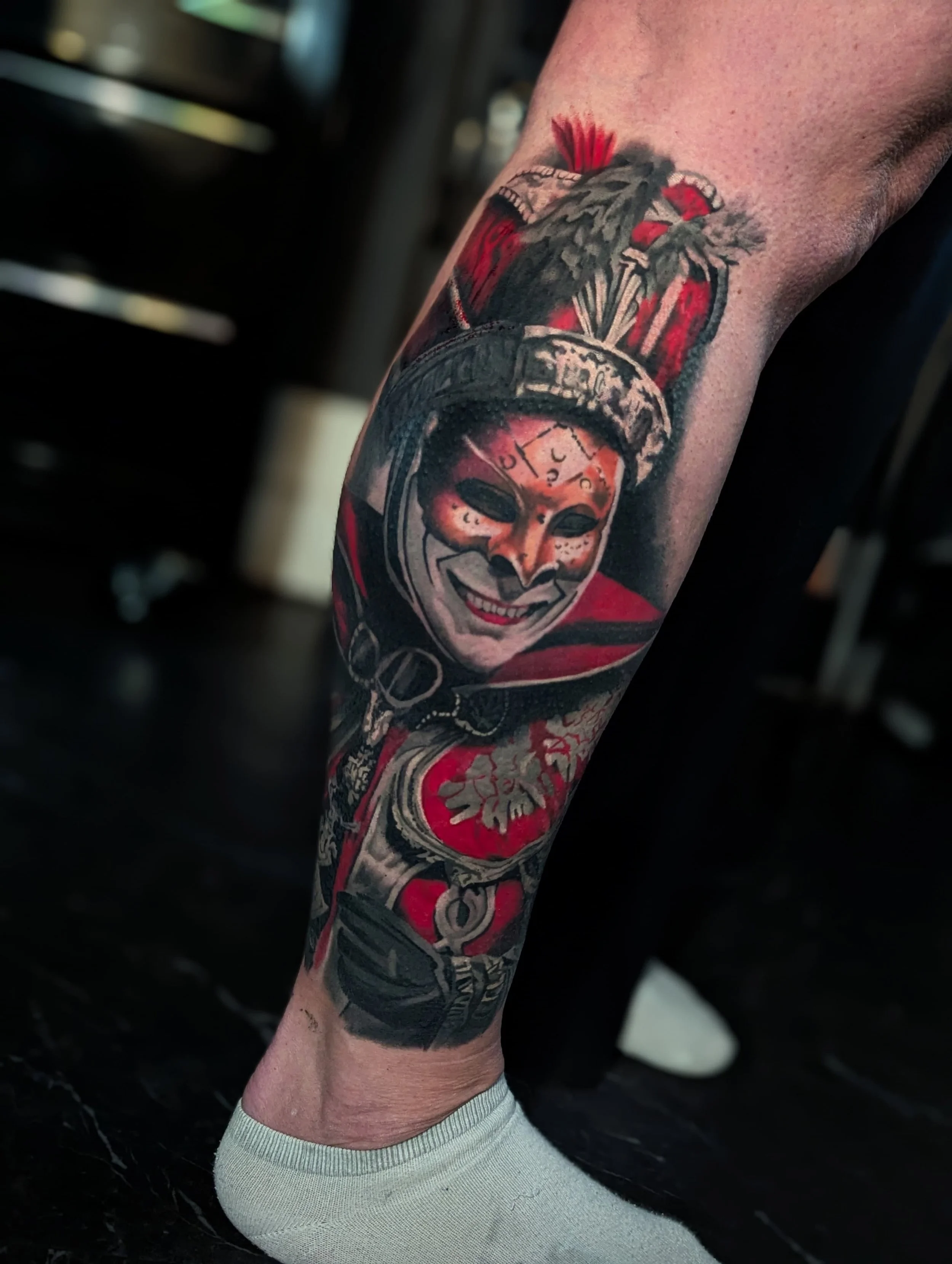 Tattoo of a smiling person wearing a creepy mask with a red and black background on a person's calf.