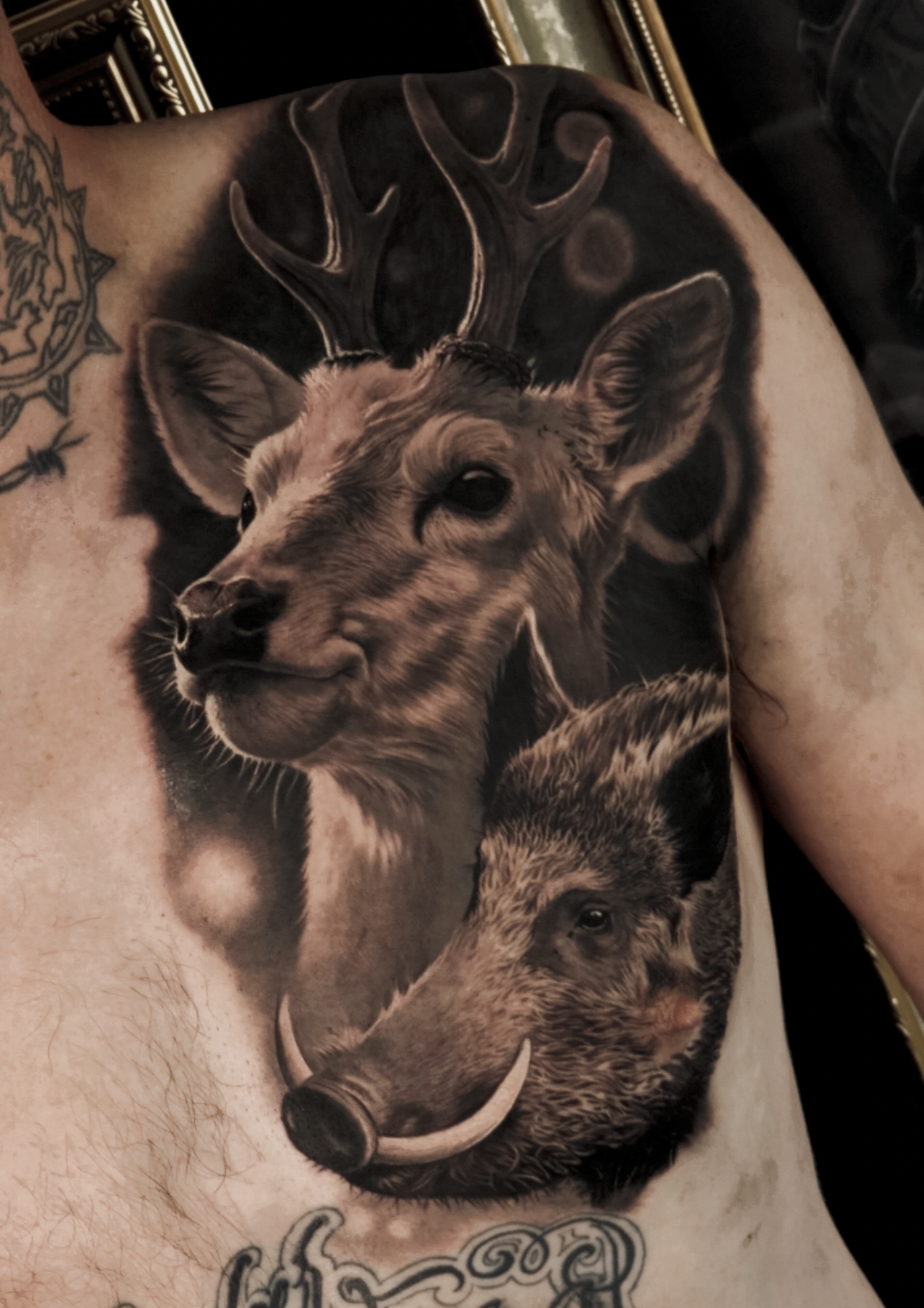 A detailed black and gray tattoo of a deer's head and antlers and a boar's head on a person's chest.