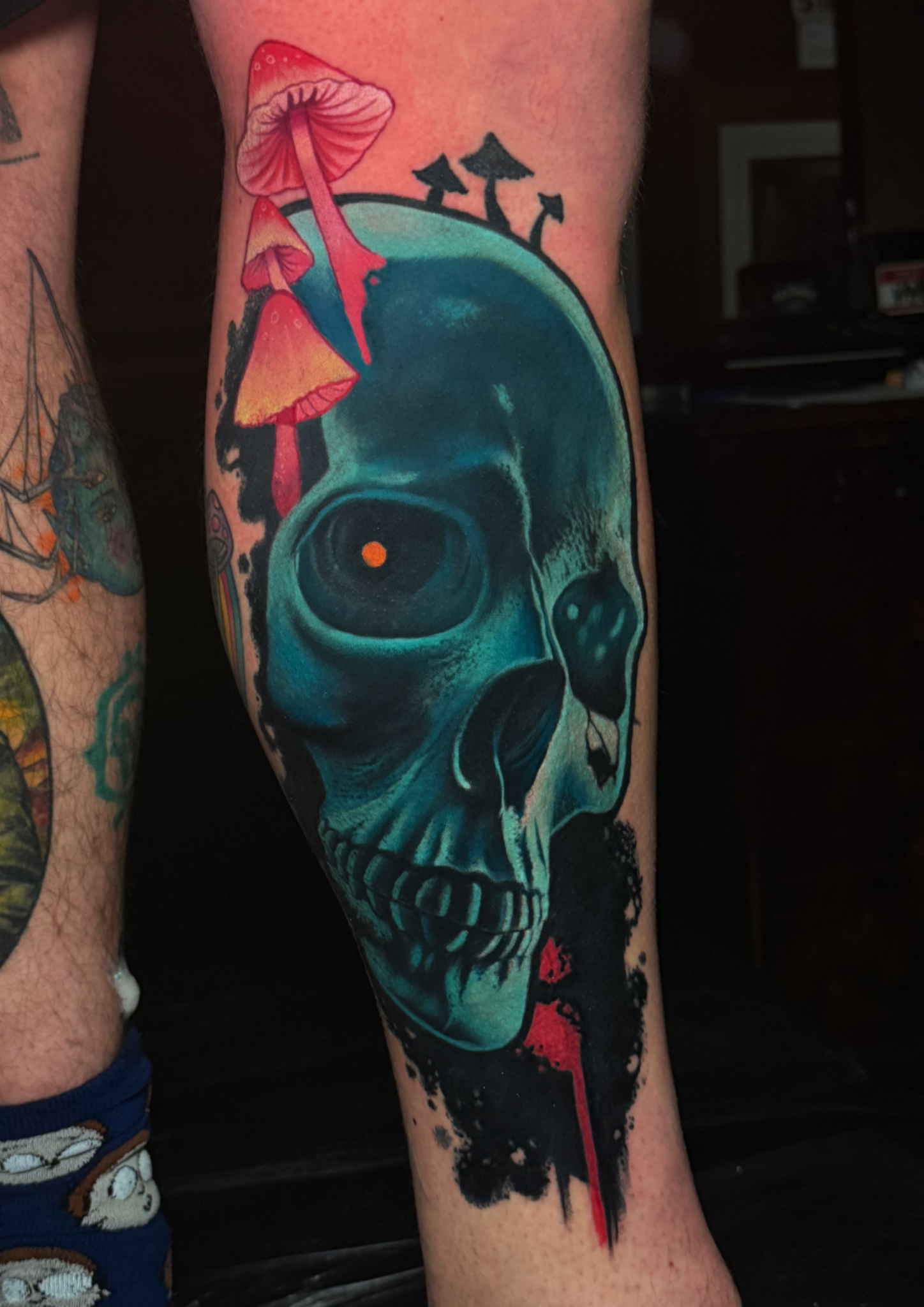 A tattoo of an alien skull in shades of blue with black and white details, featuring orange eyes and an open mouth, with colorful mushrooms and black ink splatters around it.