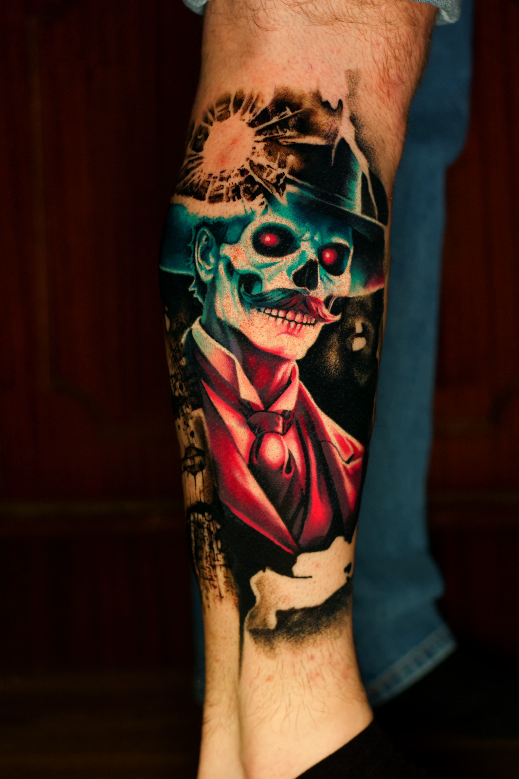 Tattoo of a skeleton wearing a suit, tie, and hat, with glowing red eyes, on a person's lower leg.