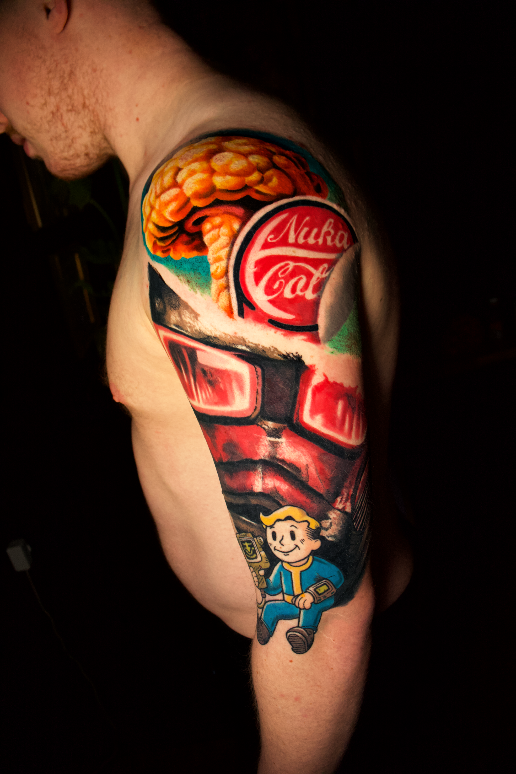 Tattoo of a mushroom cloud, a Noir Cola can, a red robot face, and a cartoon character from the Fallout video game series on a person's upper arm.