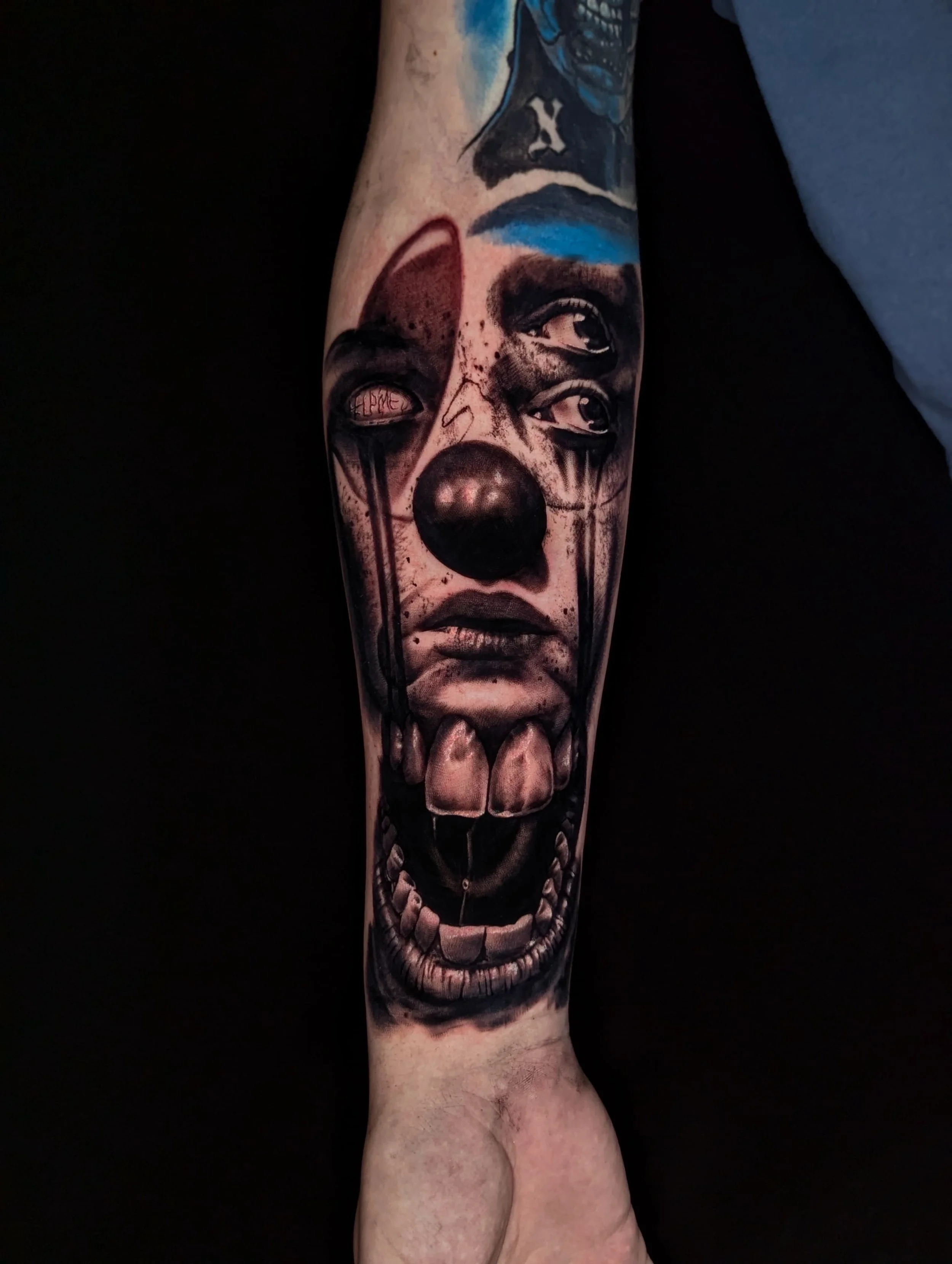 Tattoo of a clown with a black nose, open mouth showing teeth, and multiple eyes on the face, in black and gray with some color accents.
