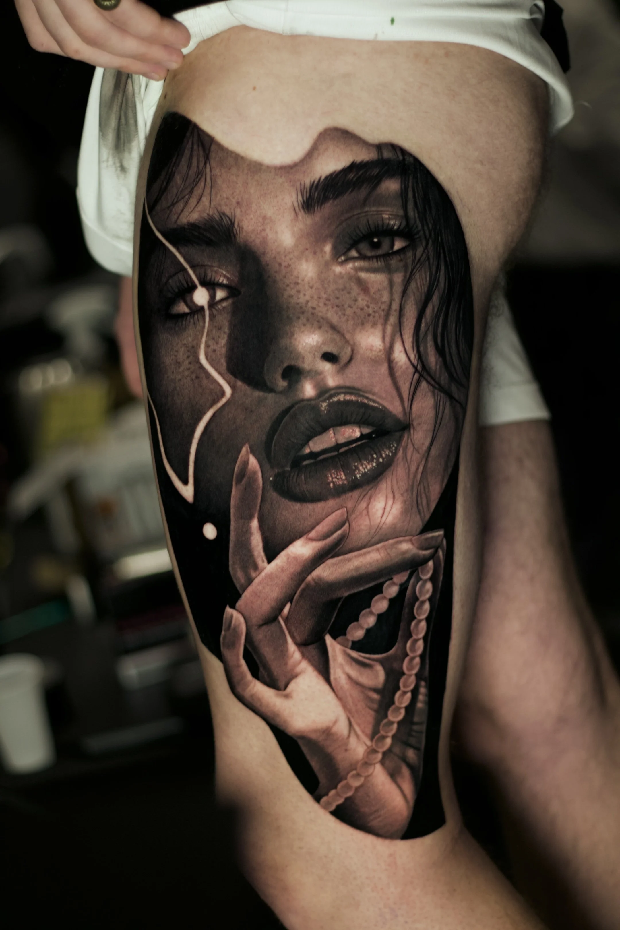 A large tattoo of a woman's face with dark makeup, freckles, and wet hair, with her finger touching her lips and a pearl necklace, covering the person's leg.