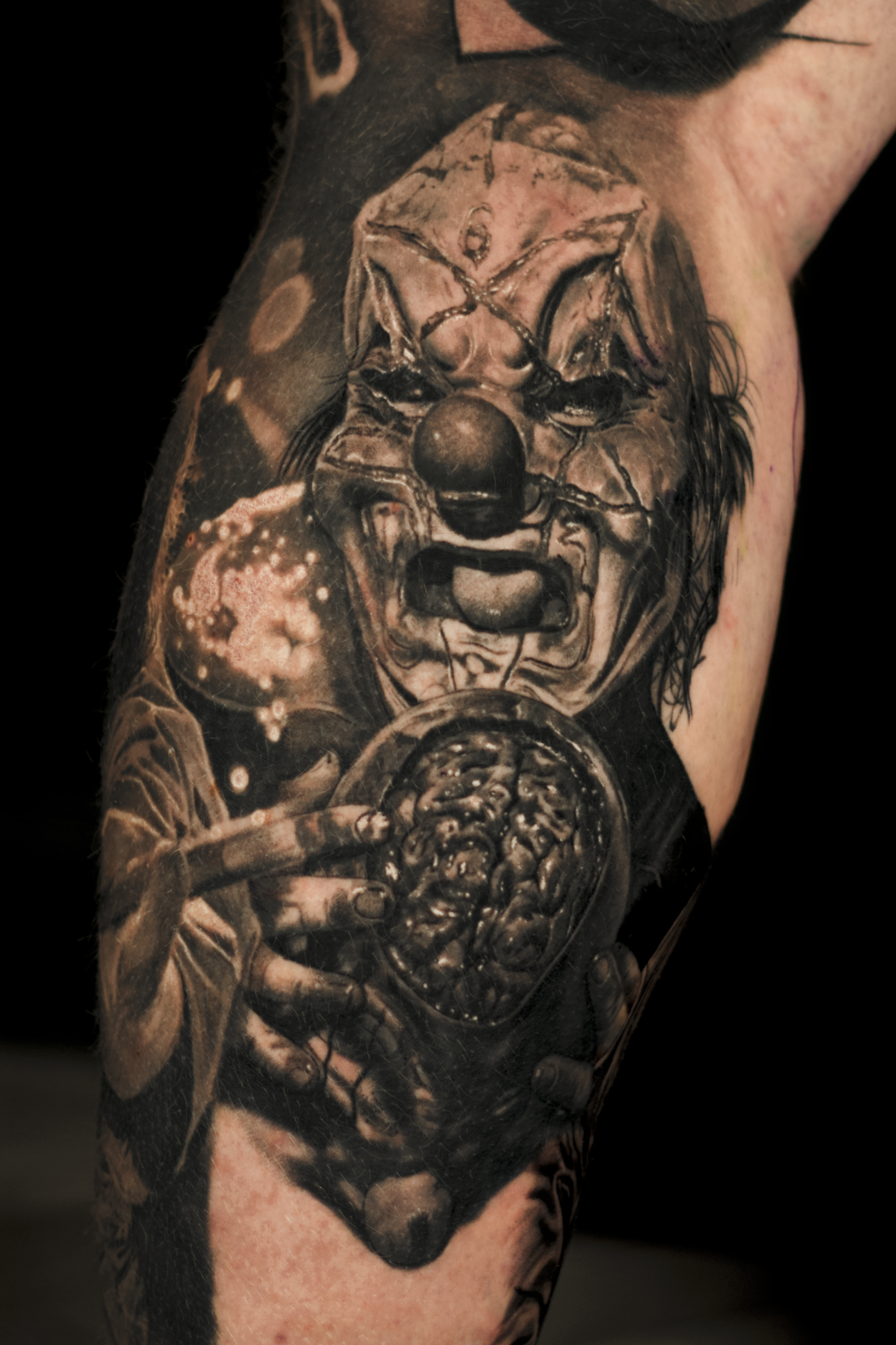Tattoo of a scary clown holding a brain in a circular dish with a dark background.