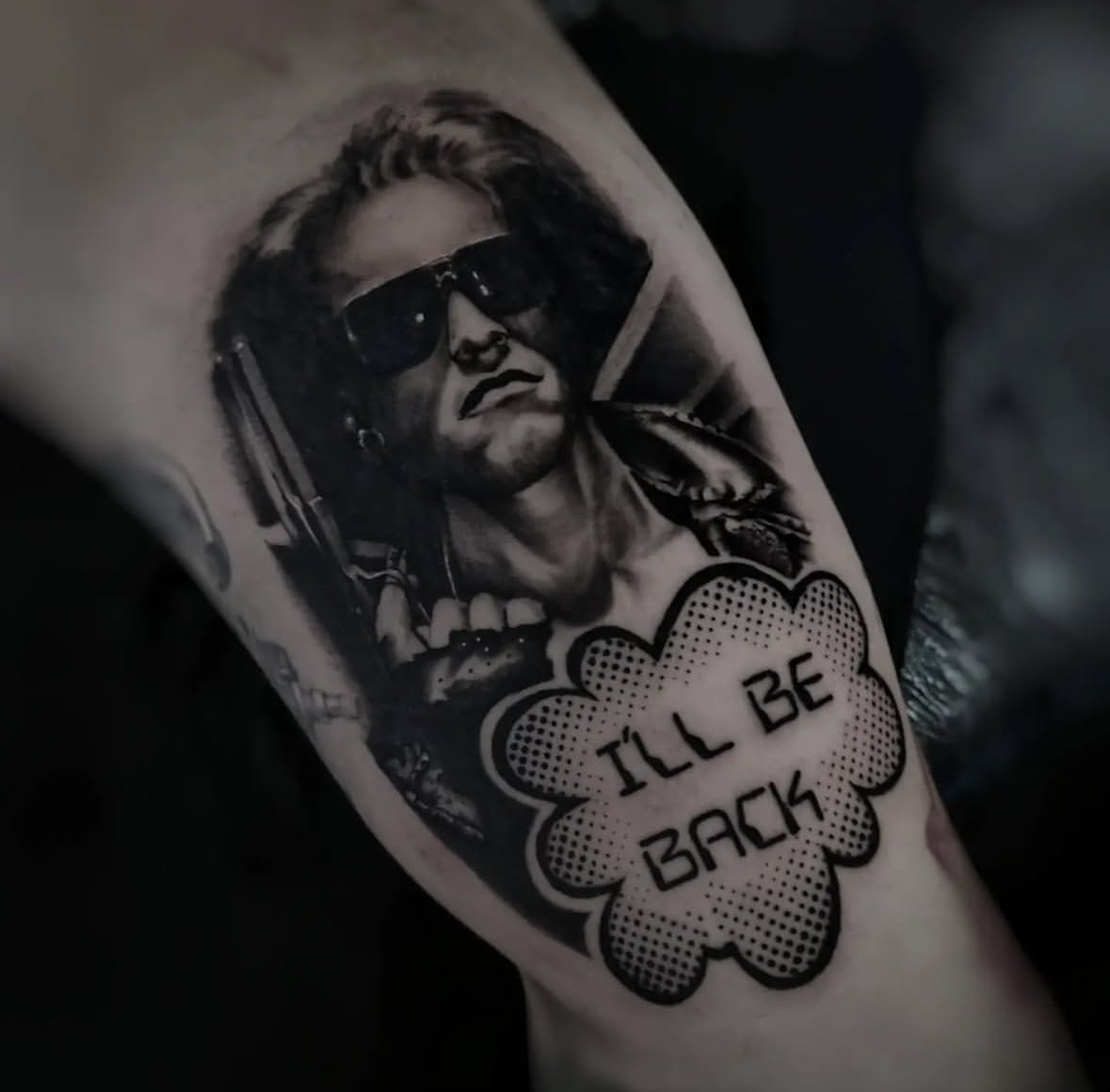 Black and white tattoo of a person wearing sunglasses and a leather jacket, holding a gun, with a speech bubble below that reads 'I'LL BE BACK'.