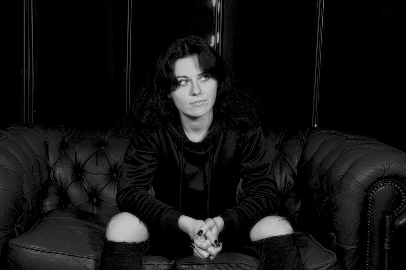 A woman with dark hair and wearing a black jacket sitting on a vintage leather couch in a room with dark walls.