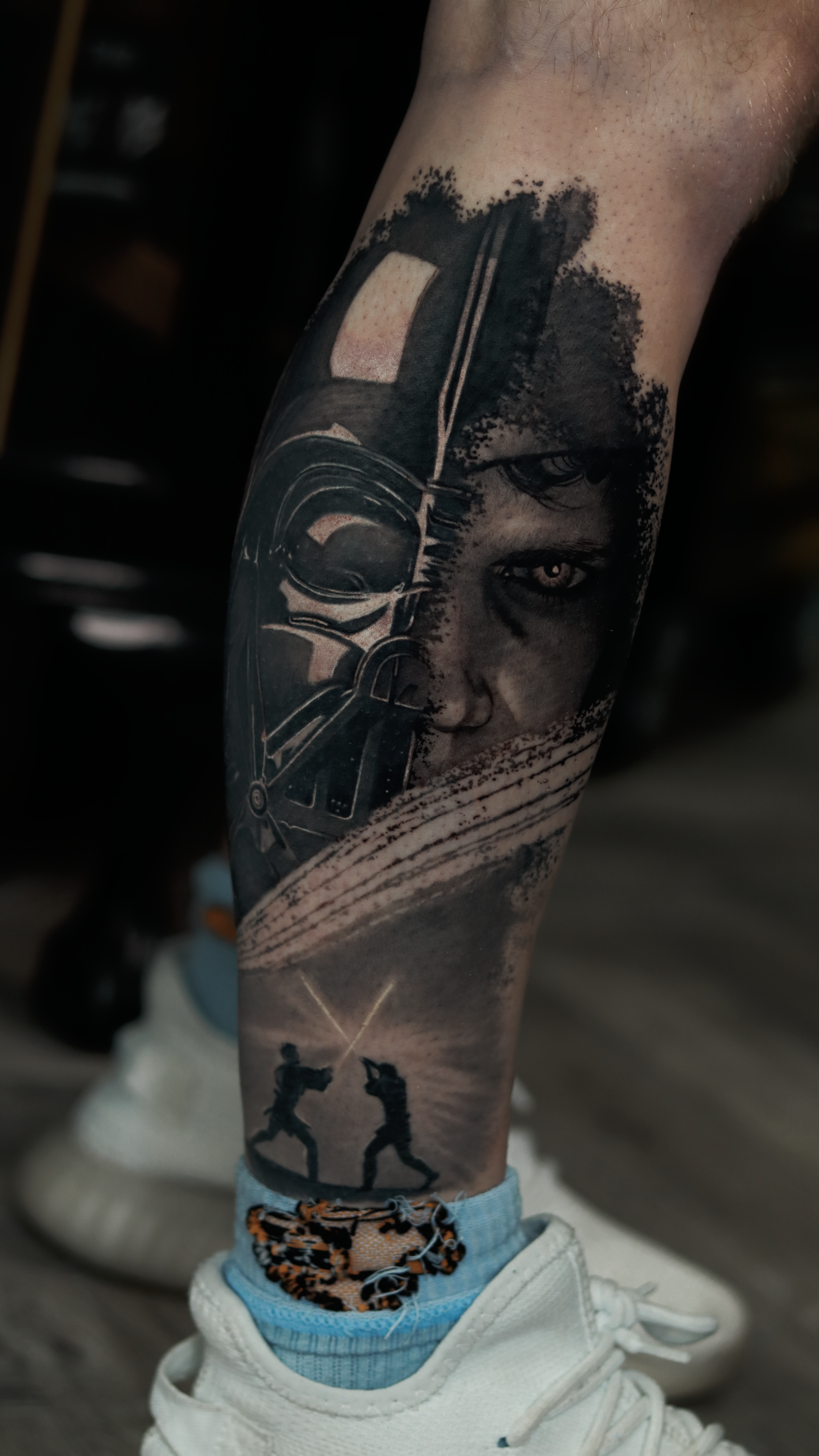 Tattoo of Darth Vader's helmet and a woman's face with intense eyes on a person's calf, with a scene of two figures fencing at sunset on the ankle.
