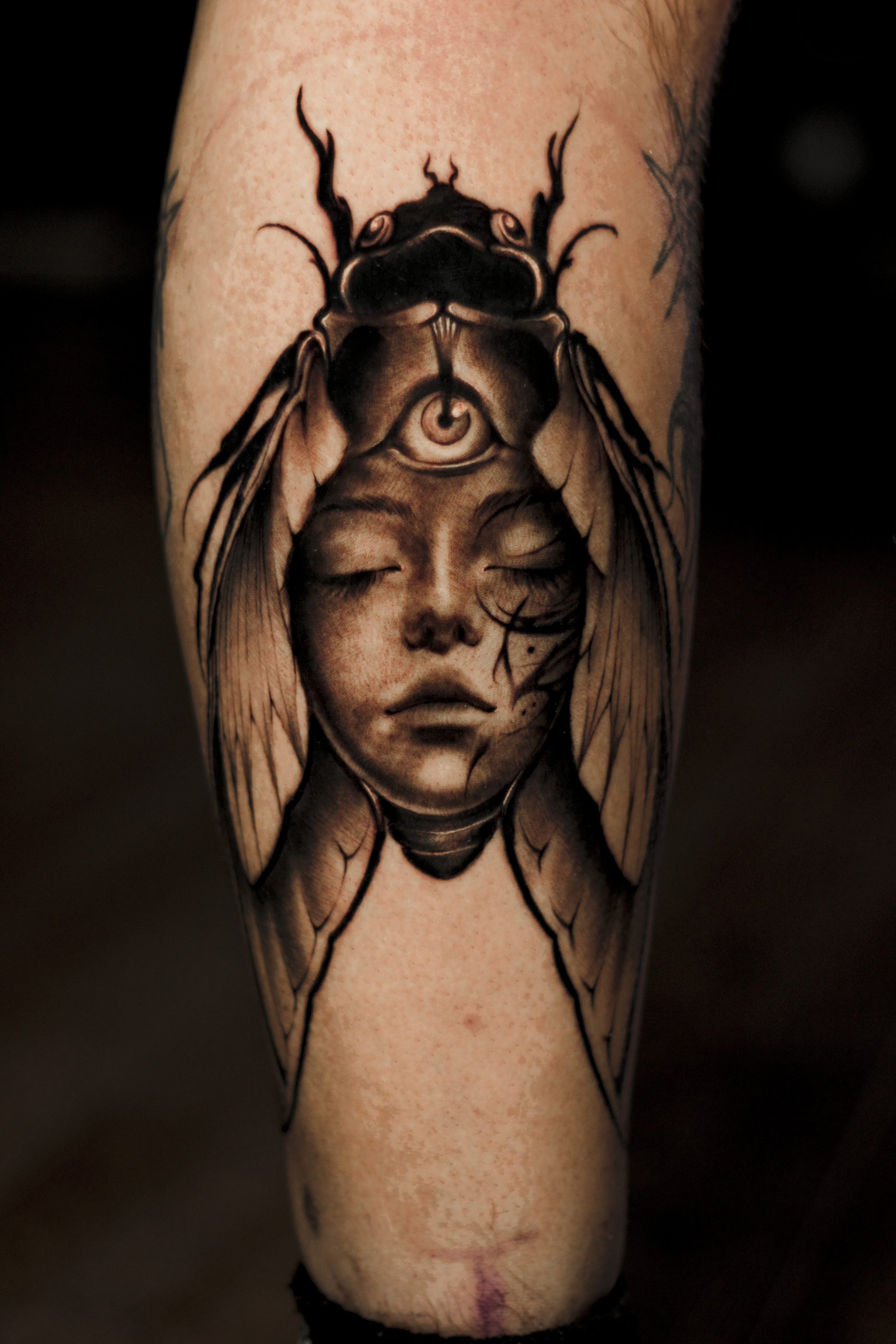 Tattoo of a woman’s face with closed eyes, combined with a large insect, possibly a beetle, with its body on top of her head, which has a third eye on its forehead.