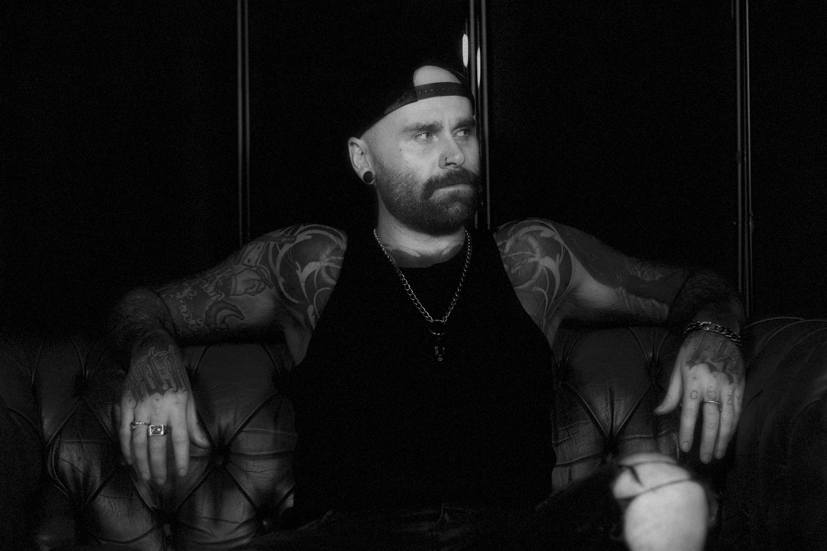 A man with a beard, tattoos, and a nose piercing wears a backwards cap and a black tank top, sitting on a leather couch in a dark room.