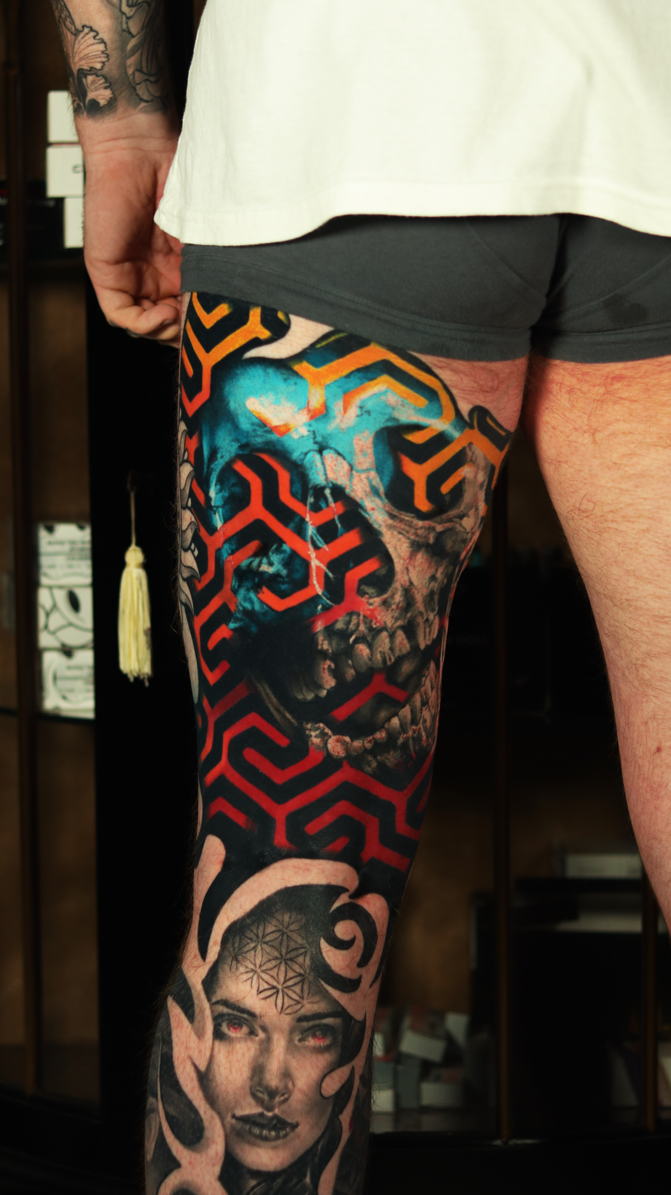 Close-up of a person's leg showcasing a detailed tattoo of a human skull and face, colorful geometric patterns, and a female portrait with red eyes and black lips on the calf, in a room with wooden shelves.