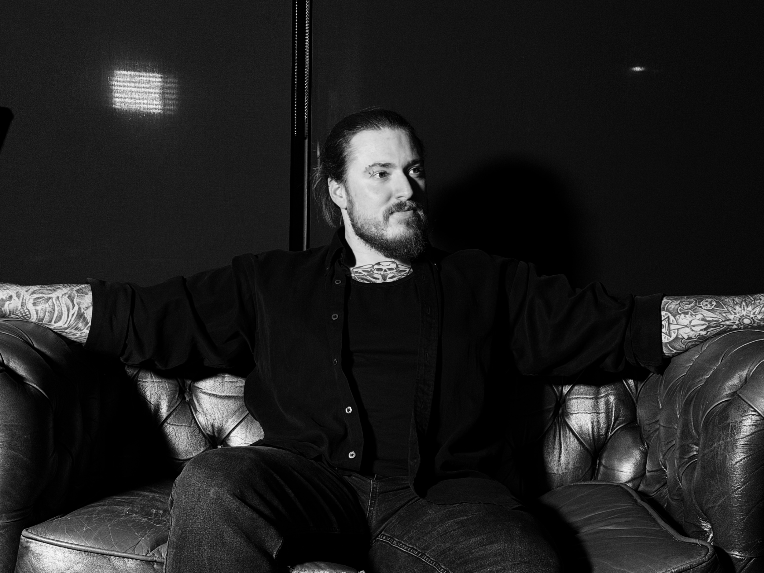 A man with long hair and a beard sitting on a leather couch with arms outstretched, against a dark background.