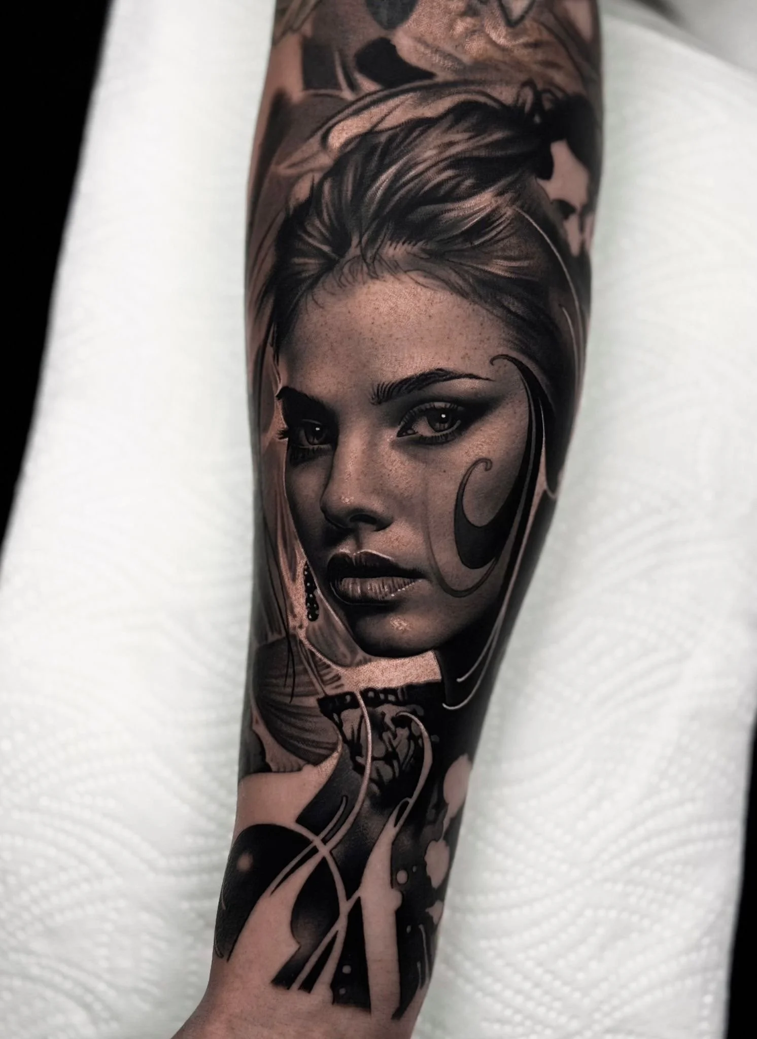 Black and gray portrait tattoo of a woman's face on a person's forearm, with detailed shading and swirling decorative elements.