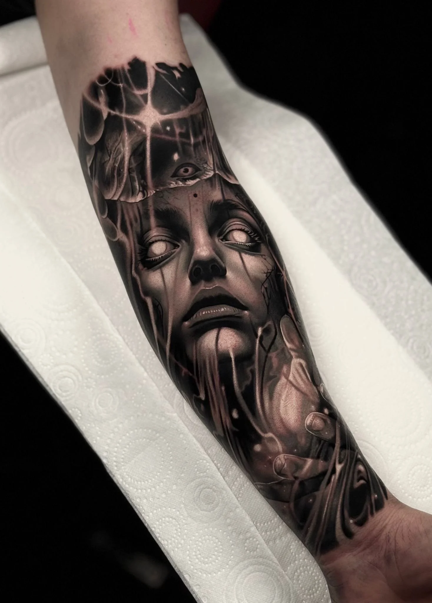 Black and gray tattoo of a woman's face with an all-seeing eye above her forehead on a person's forearm.