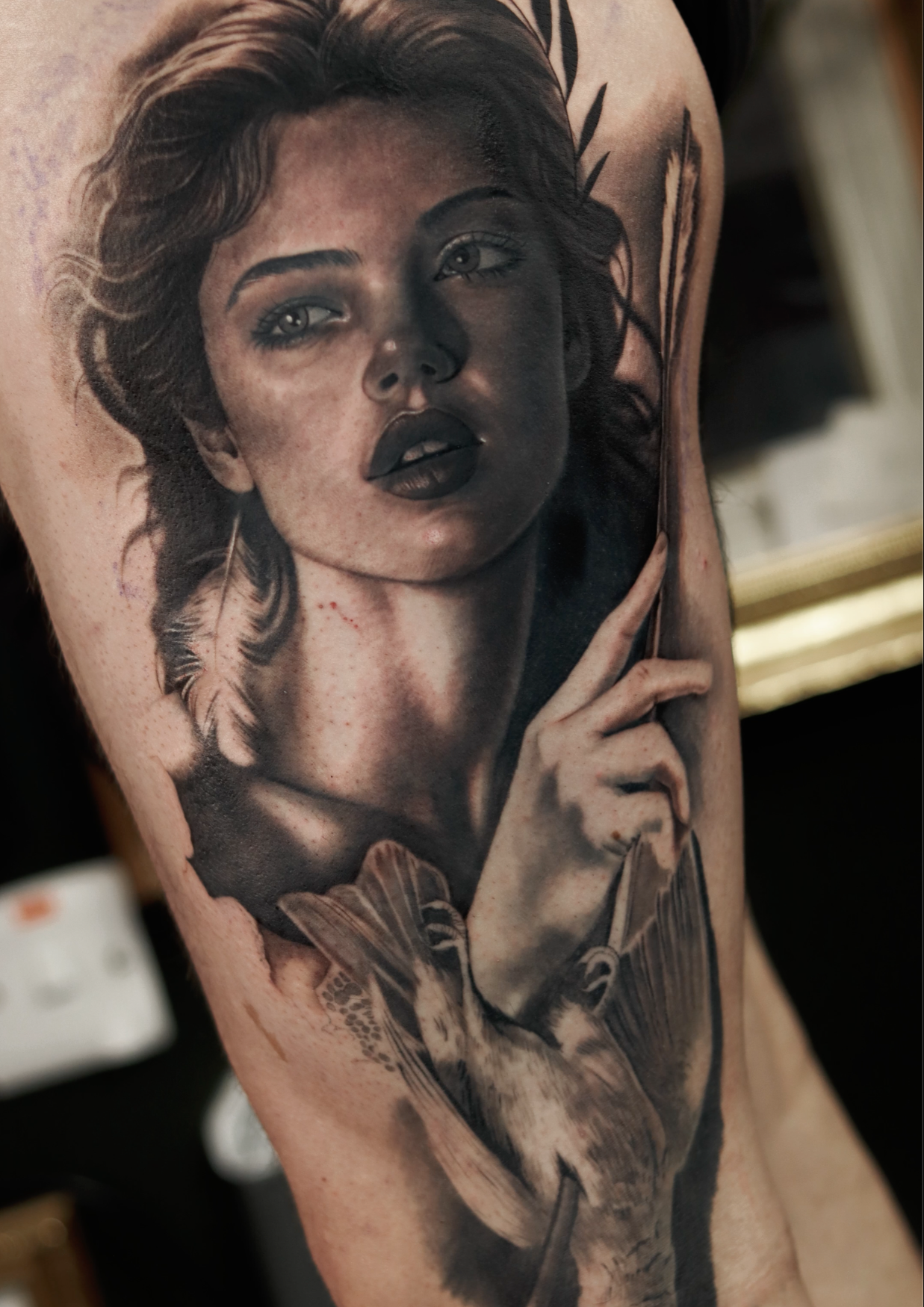 Black and gray realistic tattoo of a woman with wavy hair, intense eyes, dark lipstick, a feather earring, and a hand near her face, covering part of her neck and shoulder.