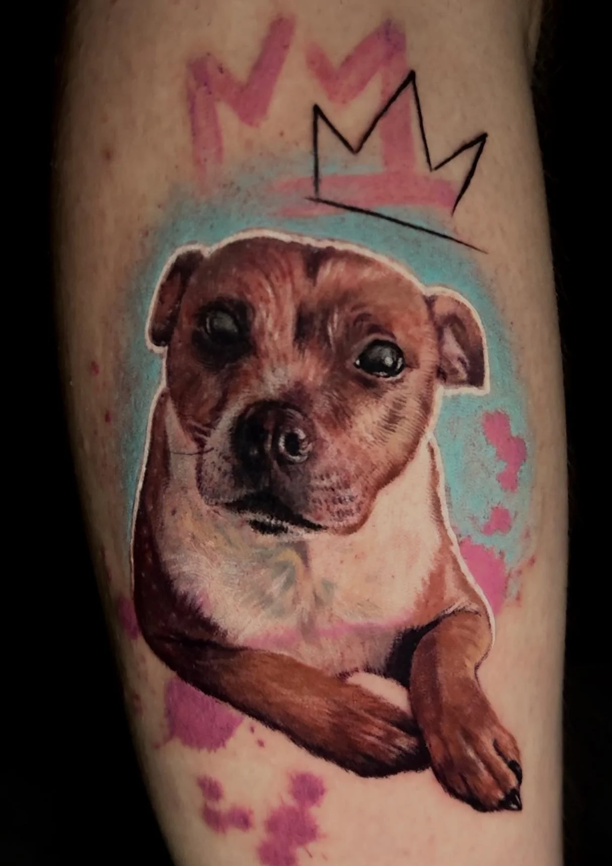 A tattoo of a realistic dog with a brown coat and expressive eyes, surrounded by pink and blue watercolor-style splashes, with a black outlined crown and pink hearts above its head.