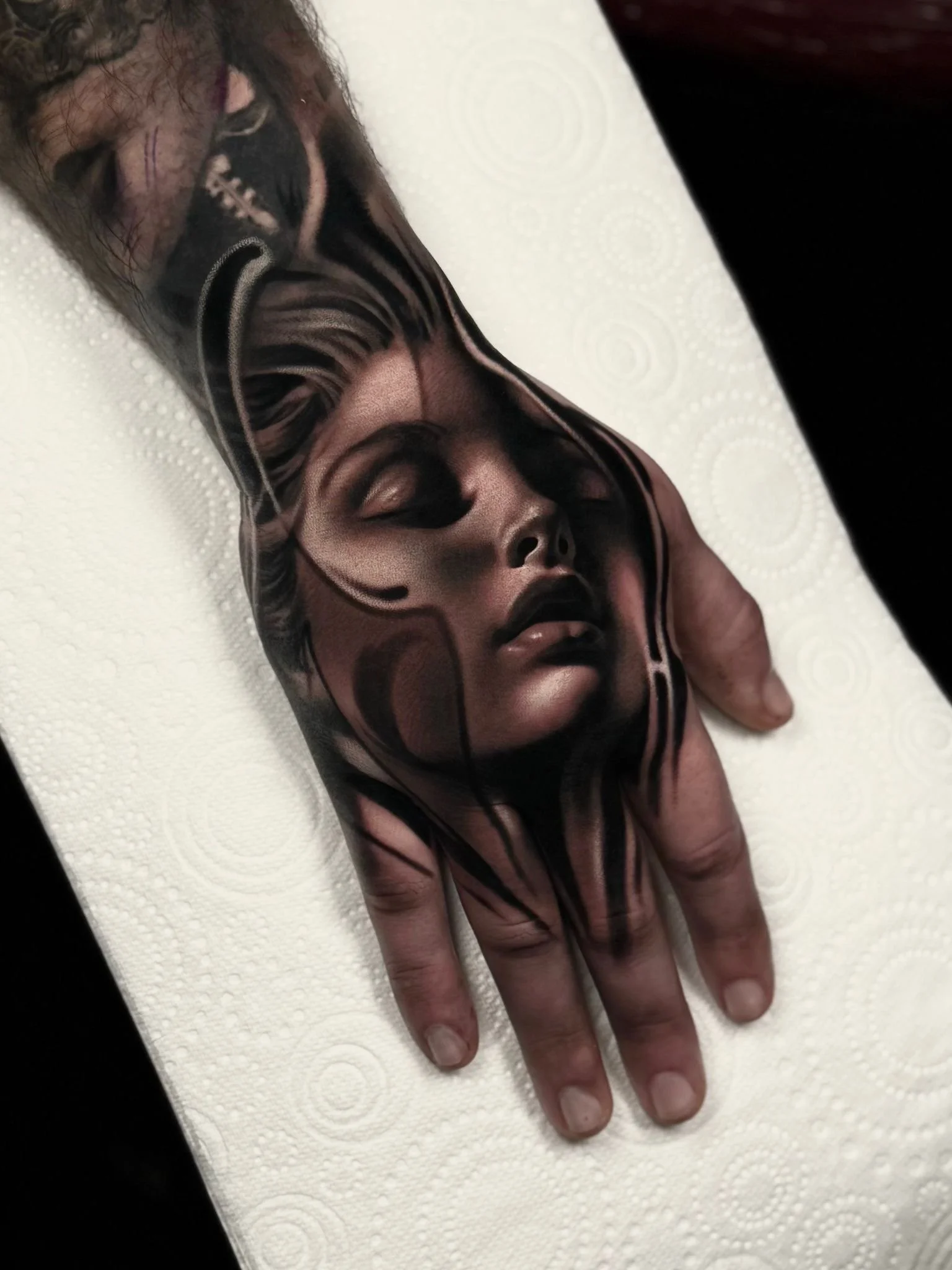 Tattoo of a woman's face with her eyes closed on a person's hand, with black and gray shading, displayed on a white textured paper towel.