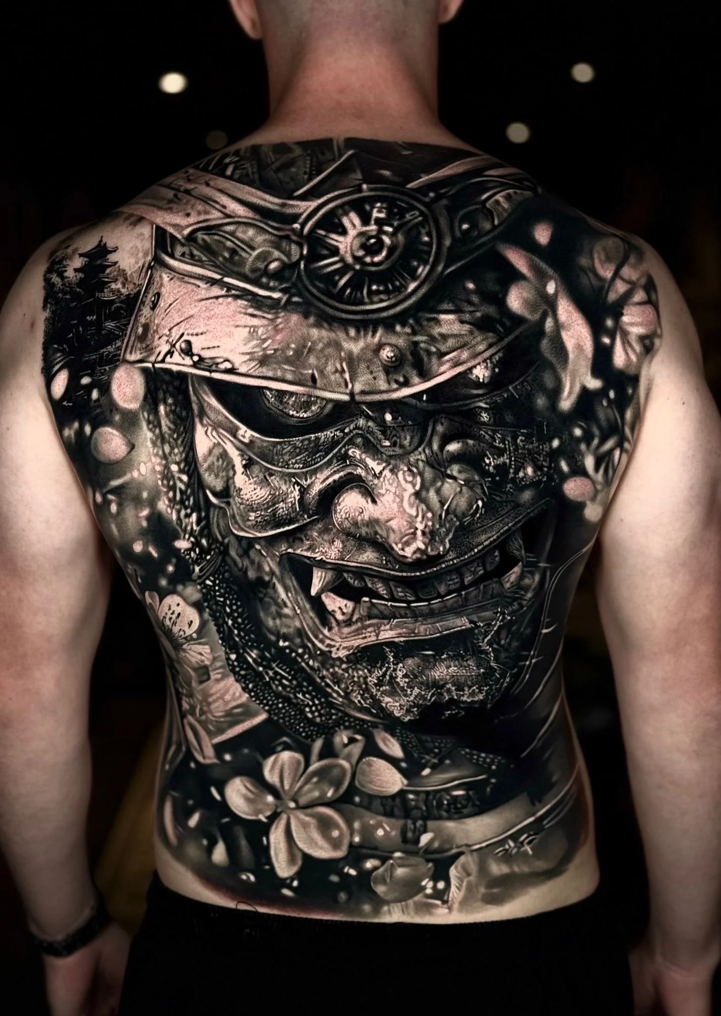 Close-up of a person's back with a large black and gray tattoo of a fierce samurai warrior's face, with detailed features, traditional armor, flowers, and intricate patterns.