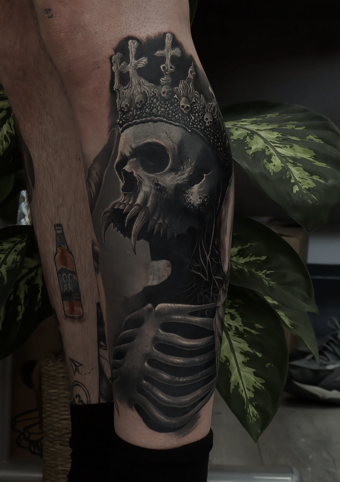 Black and gray tattoo of a skull with a crown of crosses and skulls, a skeletal hand, and a bottle labeled 'POISON'.