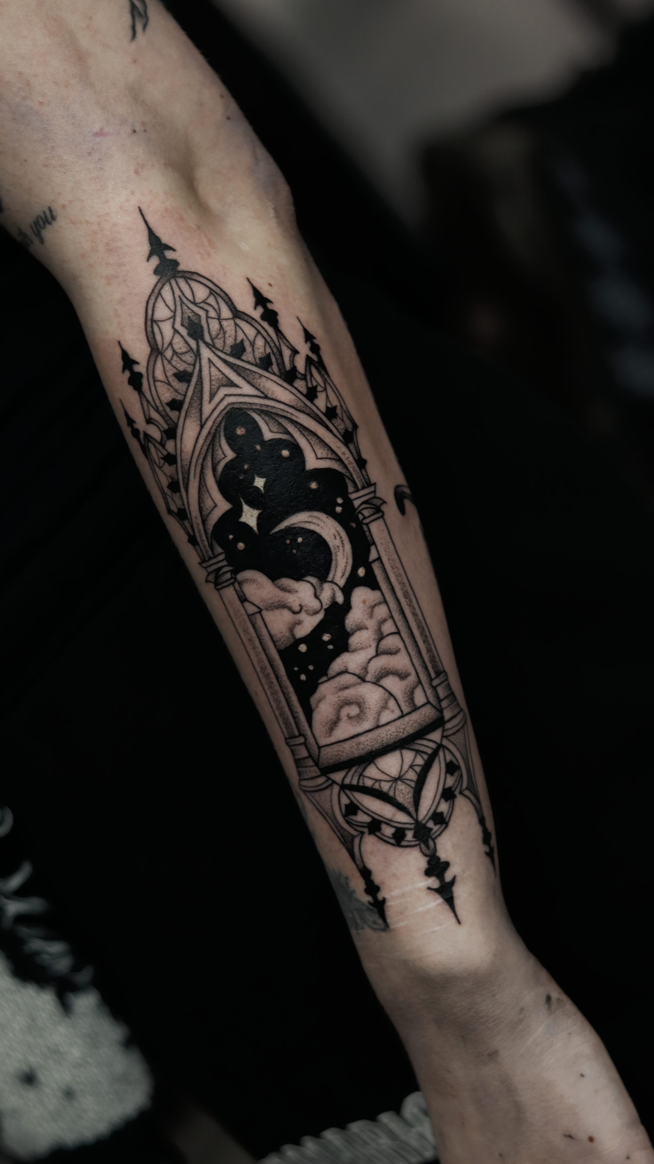 Tattoo of a gothic stained glass window design with clouds and stars on person's arm.