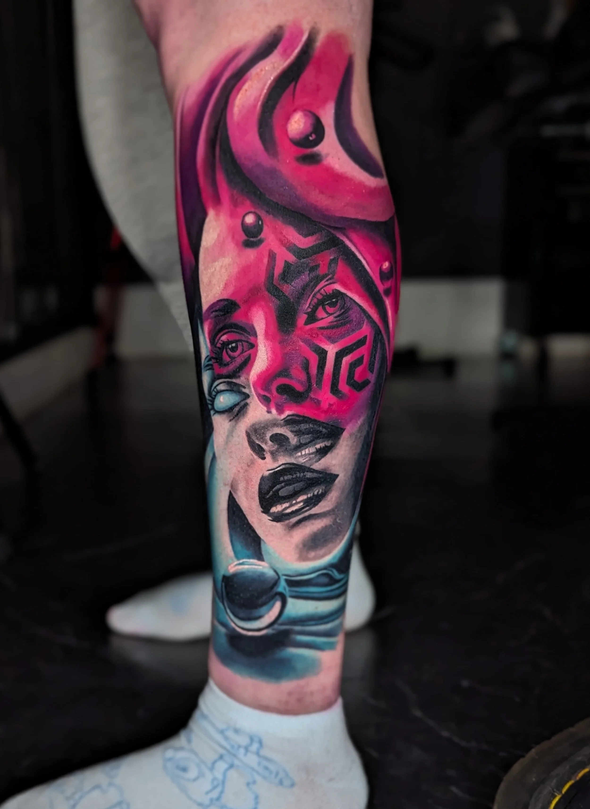 Colorful tattoo on a person's leg featuring a woman's face with bold makeup, abstract shapes, and celestial elements like planets and waves in vibrant pink, blue, and black ink.