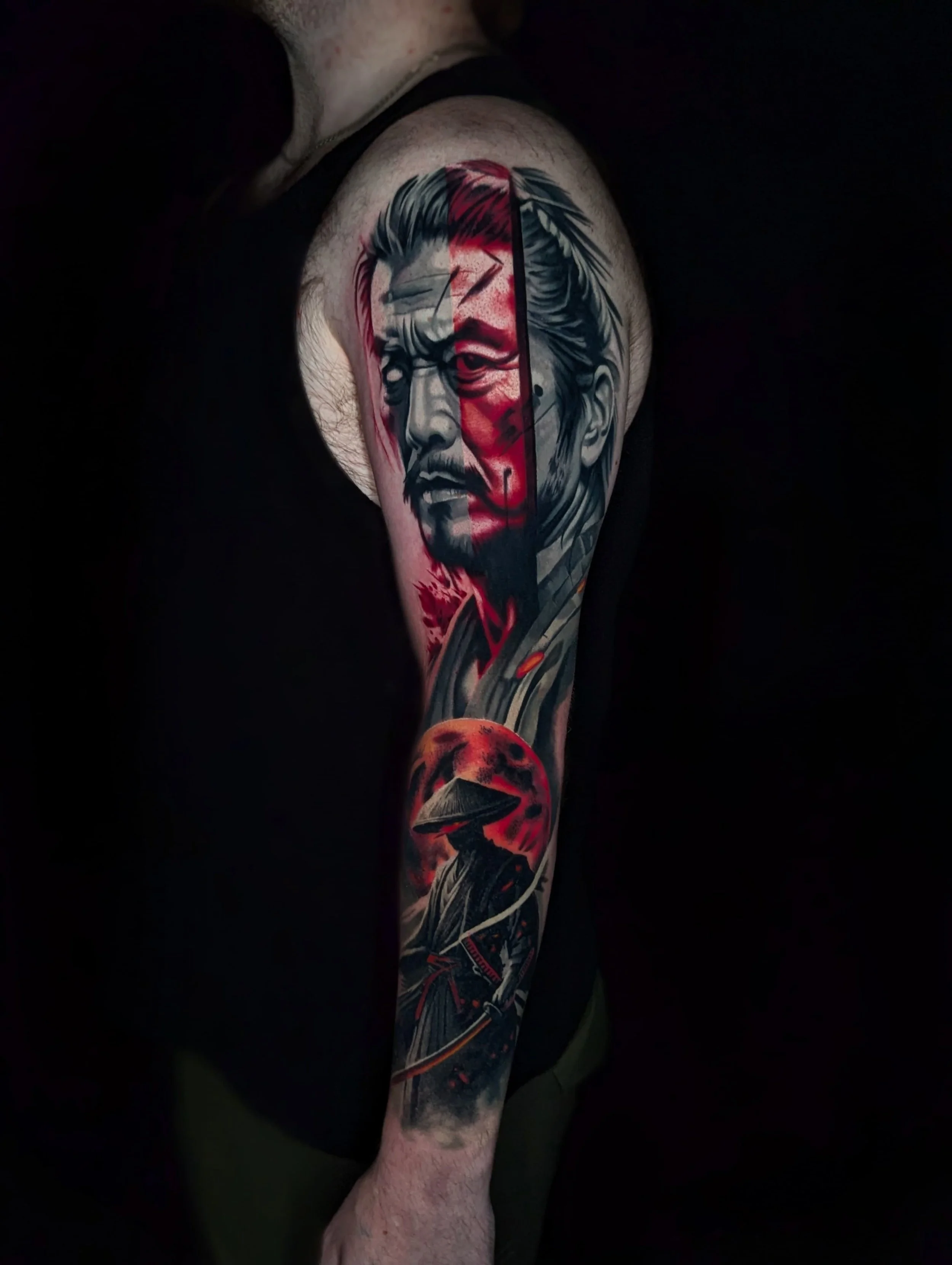 Tattoo of a warrior with a stern face and long hair on a person's upper arm, featuring a red moon in the background and a hunched figure with a conical hat and a sword at the bottom.