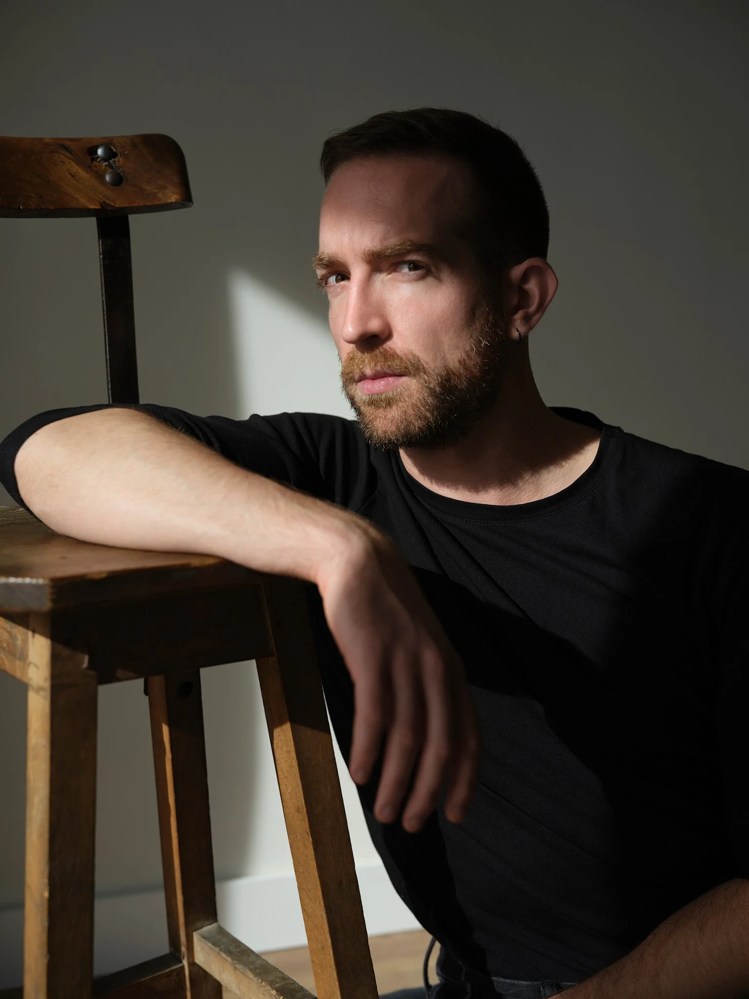 Joven con barba y auricular en tono plateado, posando con una expresión pensativa, apoyando su brazo en una silla de madera, luz natural iluminando su rostro, fondo neutro.