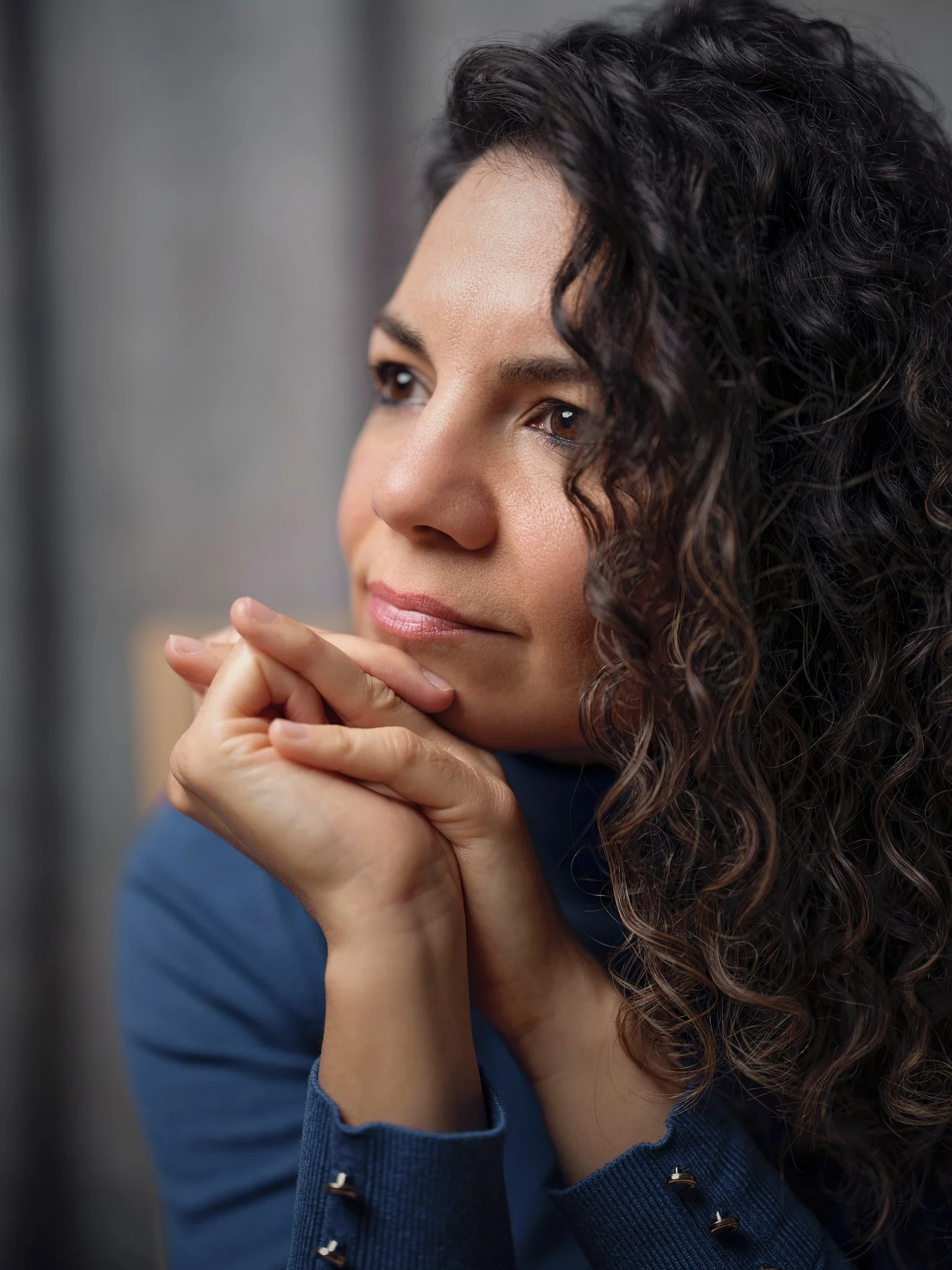 Mujer con cabello rizado mirando pensativamente, con las manos juntas apoyadas en su rostro, ropa azul.