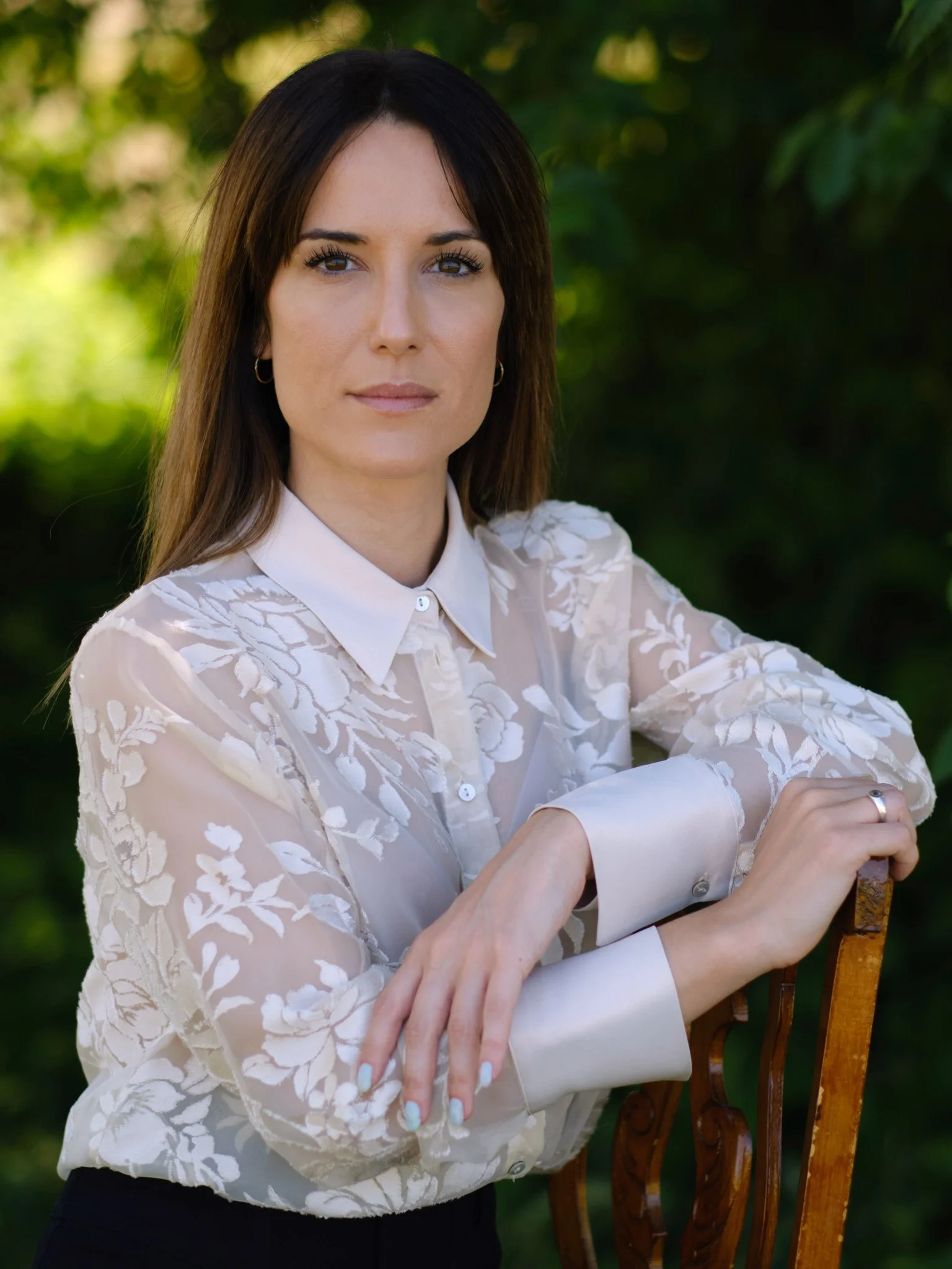 Una mujer de cabello largo y oscuro, con expresión seria, viste una blusa de manga larga translúcida con bordados blancos sobre fondo claro, posando al aire libre con fondo de vegetación.