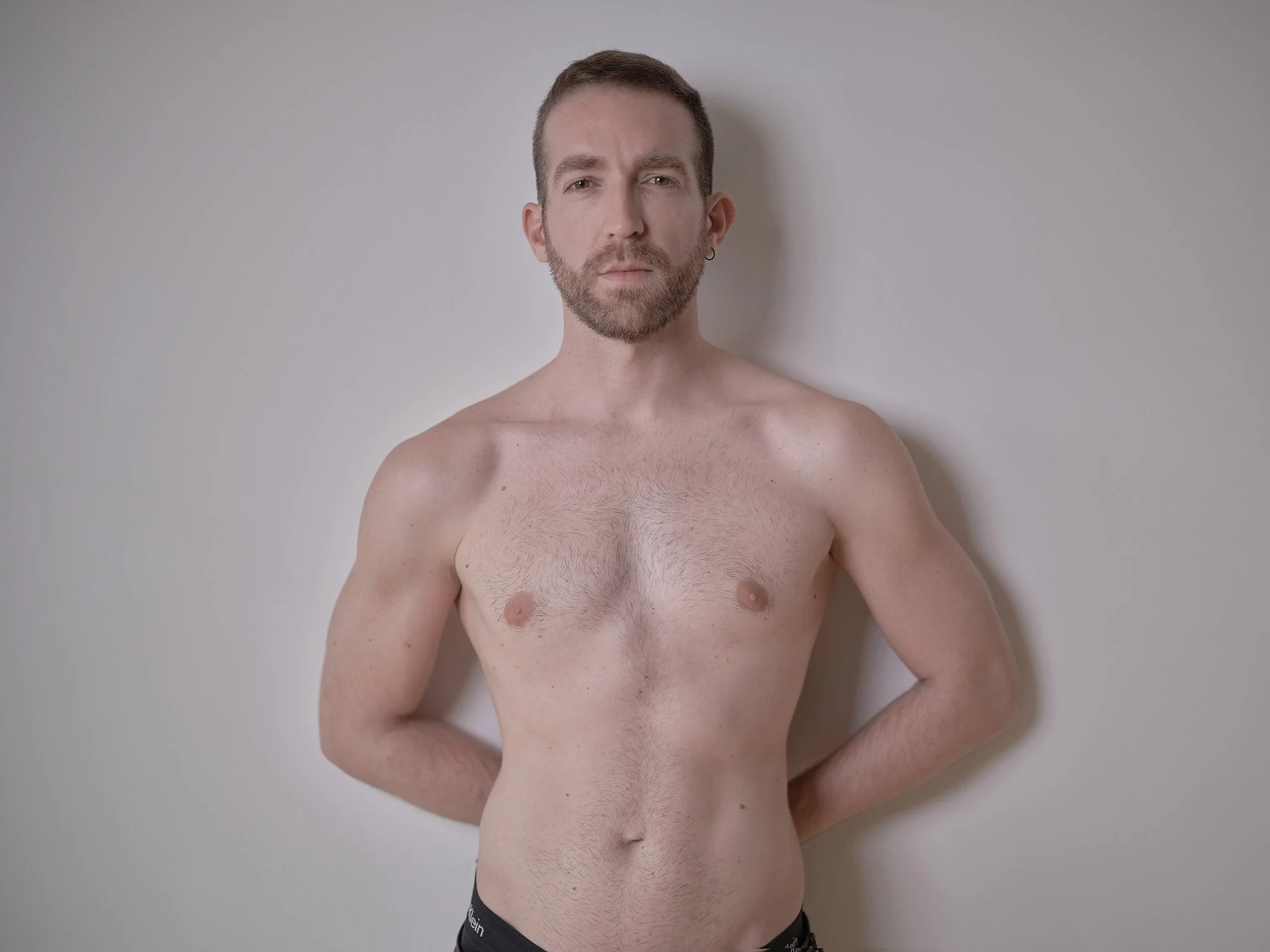 Hombre sin camisa con barba, de cabello corto, posando contra una pared blanca