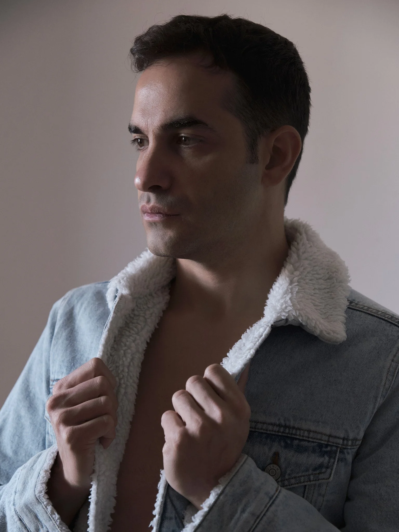 Joven con chaqueta de mezclilla con forro de felpa, mirando hacia la izquierda, apoyada en una pared de color neutro.