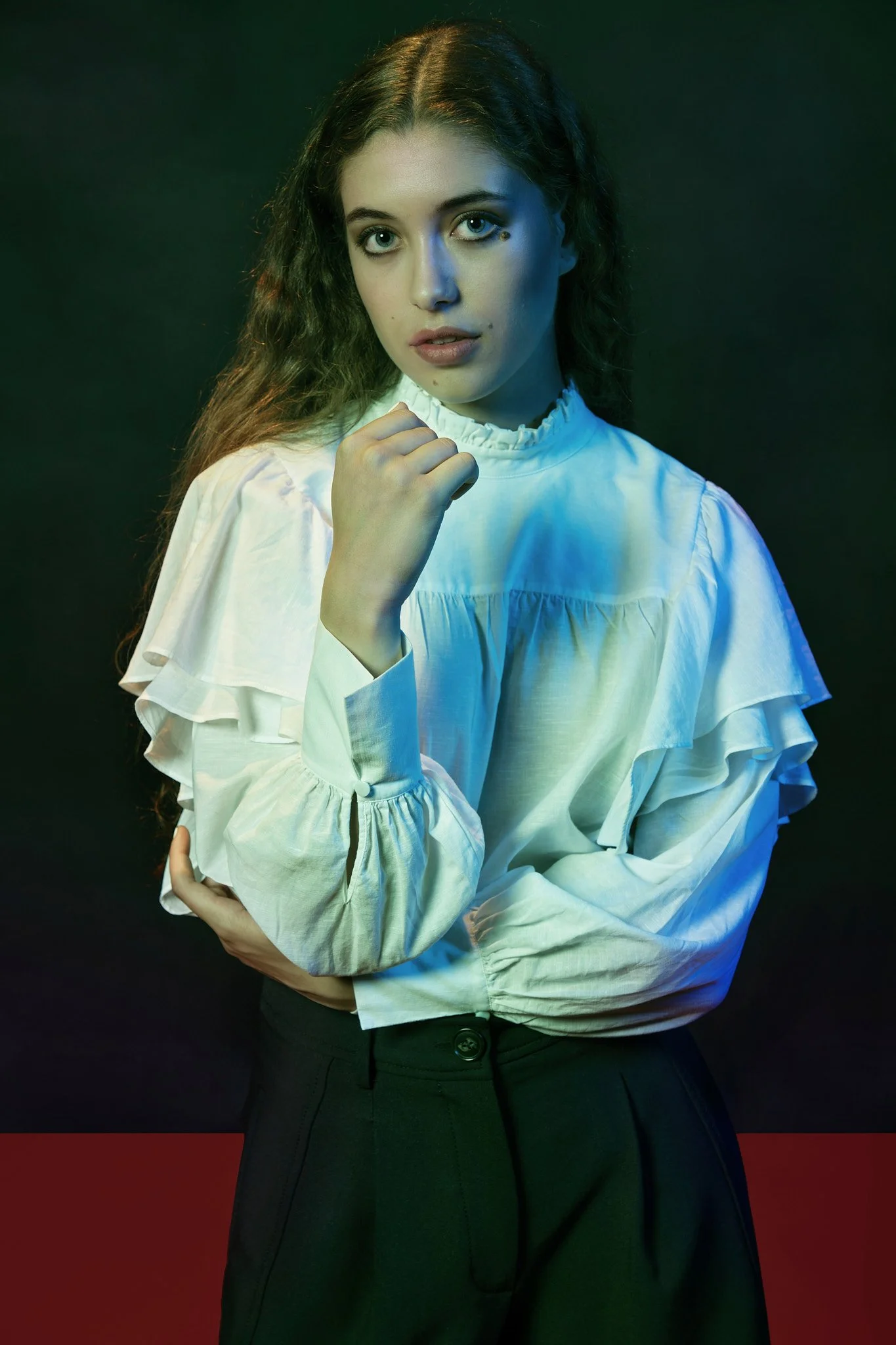 Retrato de una mujer con cabello rizado y tez clara, vistiendo blusa blanca de manga larga con volante en los hombros, en un fondo oscuro con iluminación de tonos fríos y cálidos.