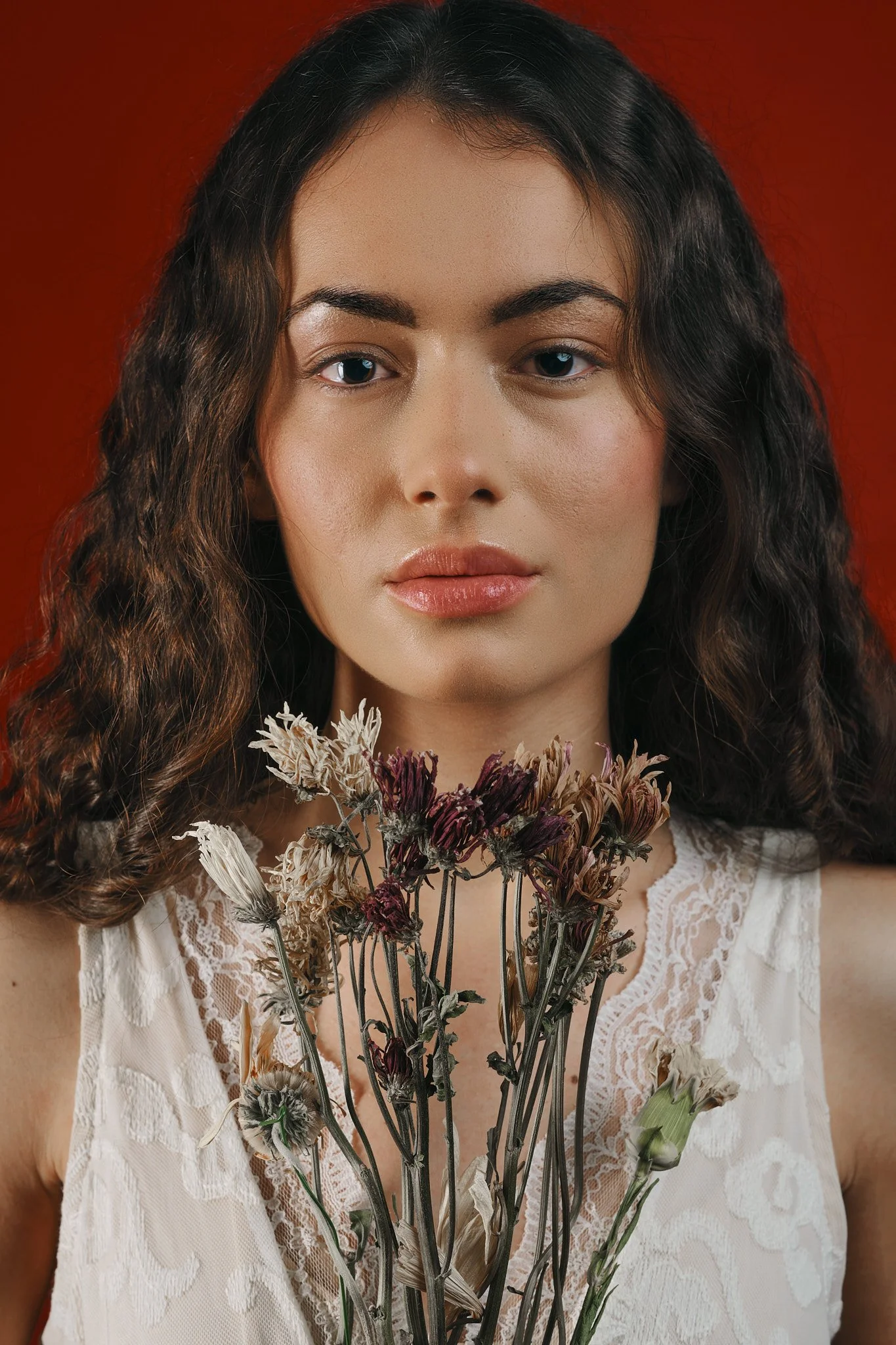 Retrato de una mujer con cabello rizado sosteniendo flores secas en el pecho, lleva blusa de encaje blanca, fondo rojo.