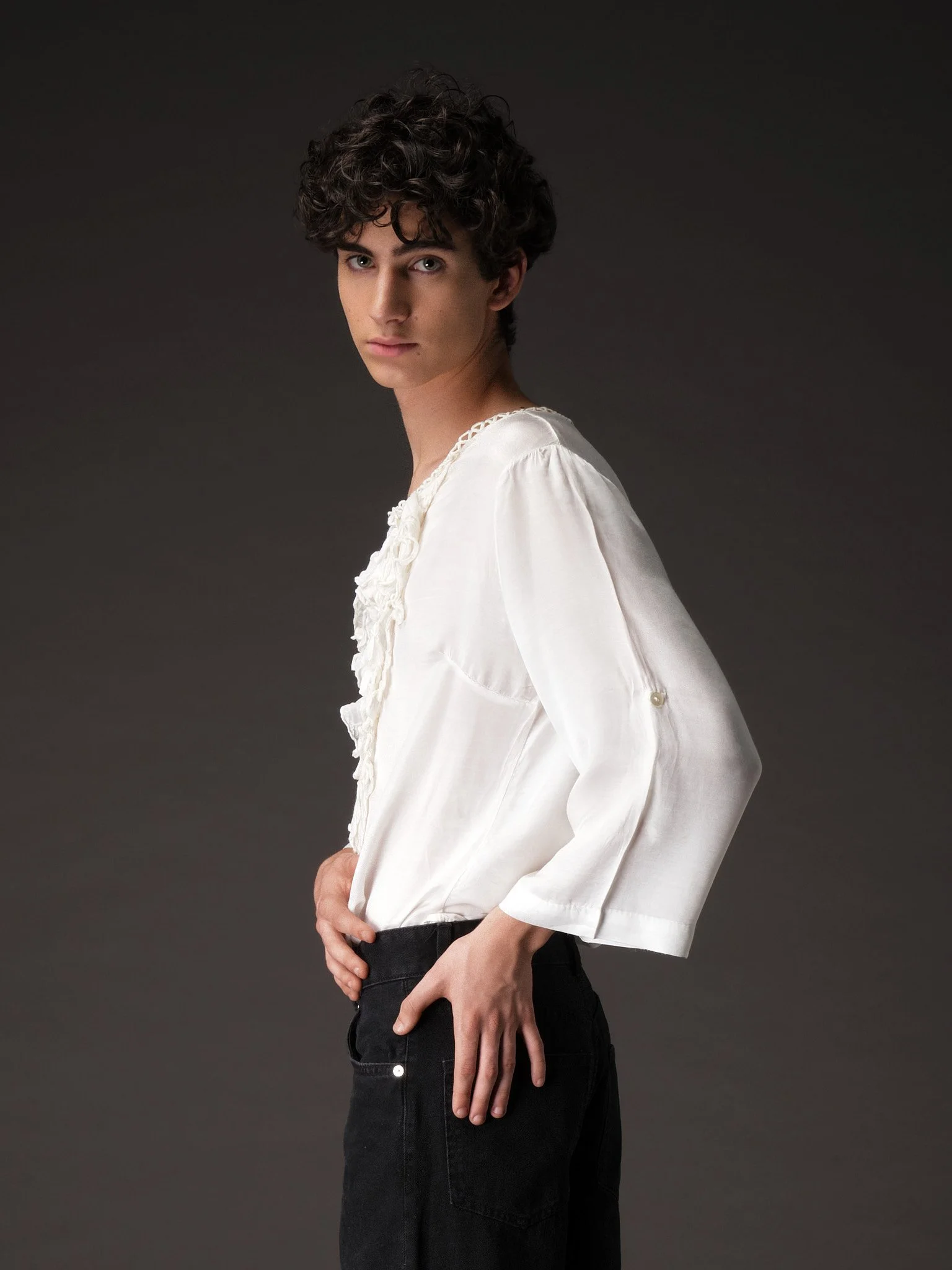 Joven con cabello rizado oscuro, vistiendo blusa blanca de manga larga y pantalones negros, posando de perfil con fondo gris oscuro.