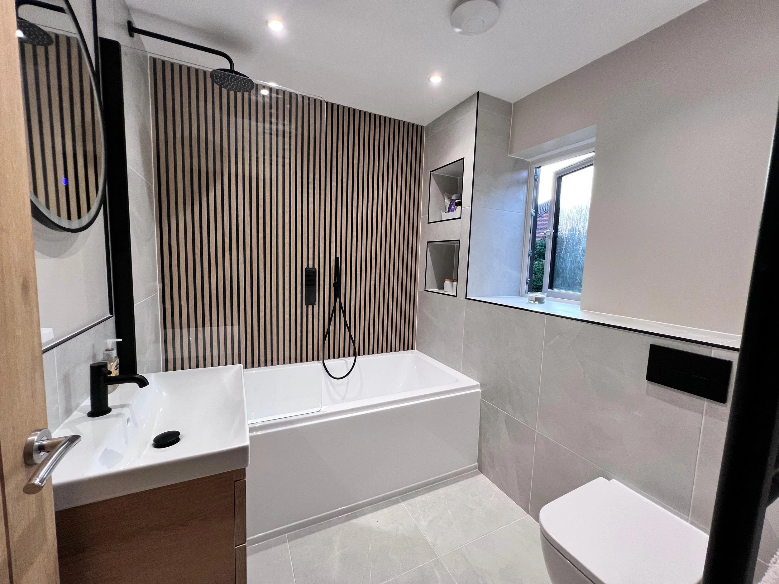 White bath against wooden background with black taps and shower head. White sink on wooden cabinet with circle mirror above