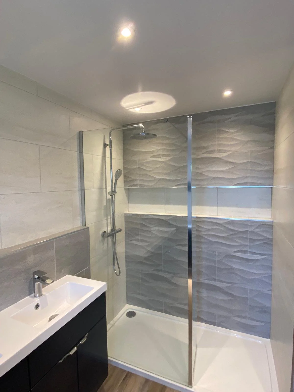 Modern bathroom with a glass shower enclosure, textured gray tiles on the shower wall, a white shower tray, a small black vanity with a white sink, and wood-look flooring.
