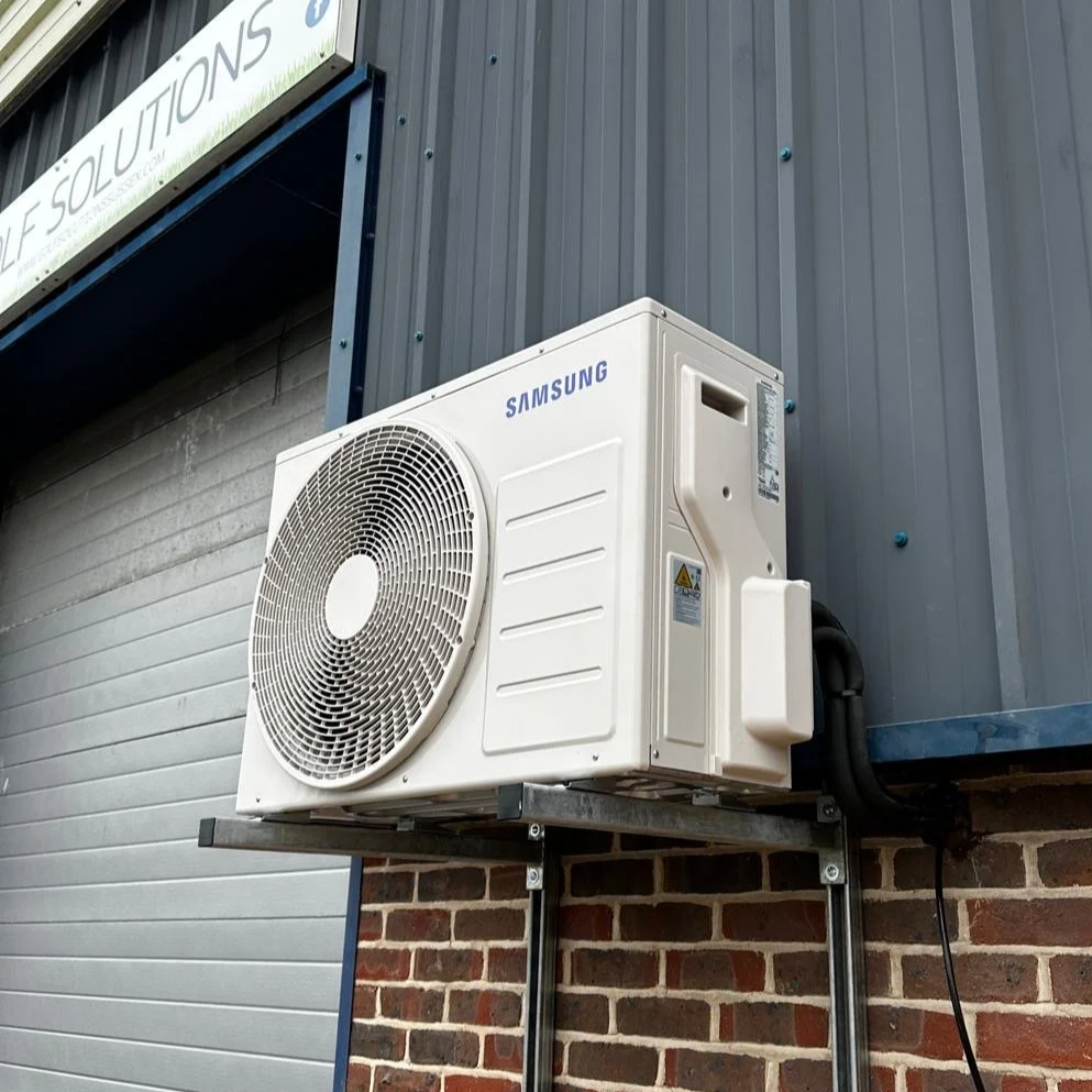 Samsung air conditioning unit installed outside on a metal stand next to a brick wall and gray siding.